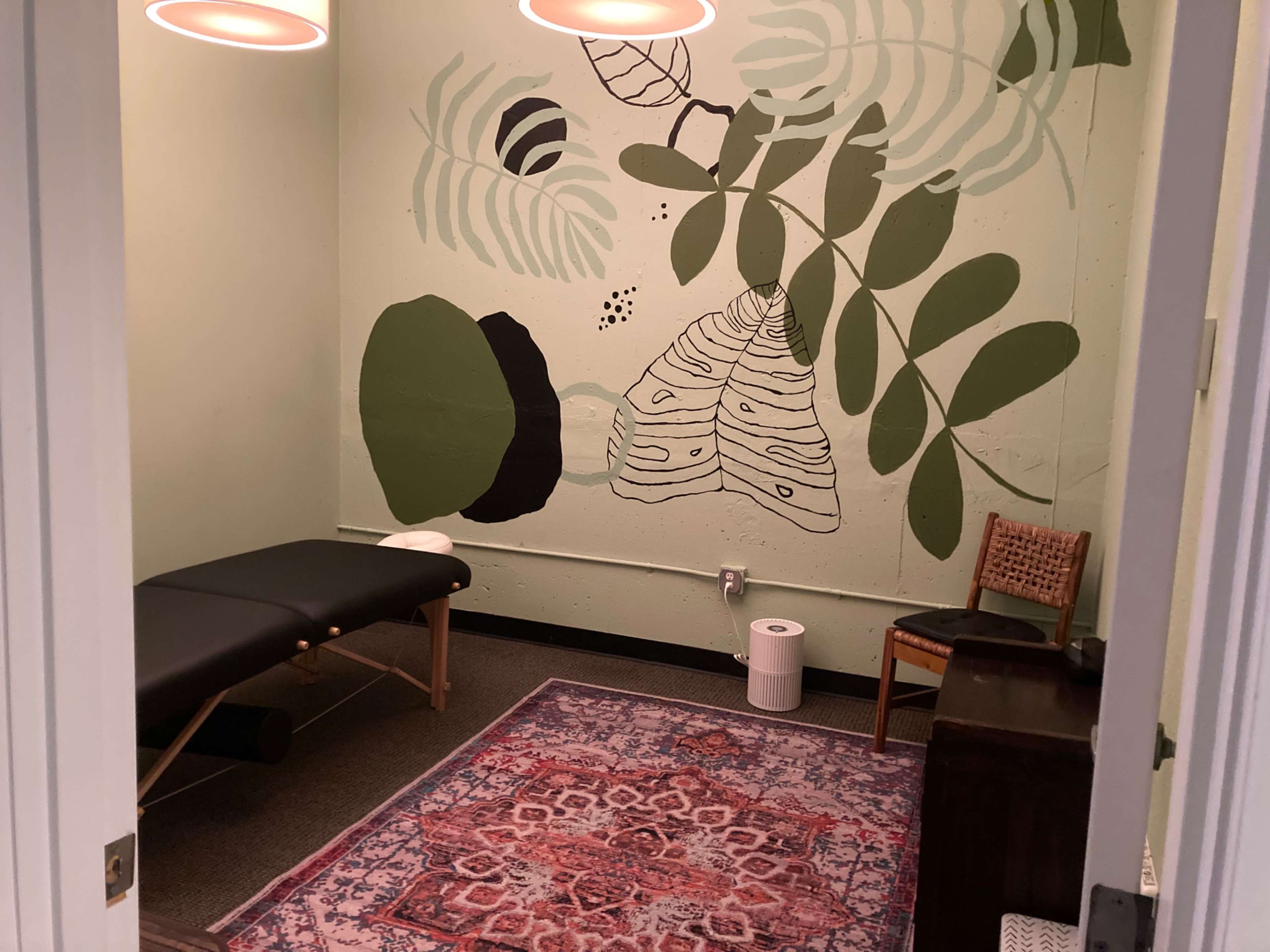 A small room with a massage table, a patterned rug, and a mural of plants on the wall.