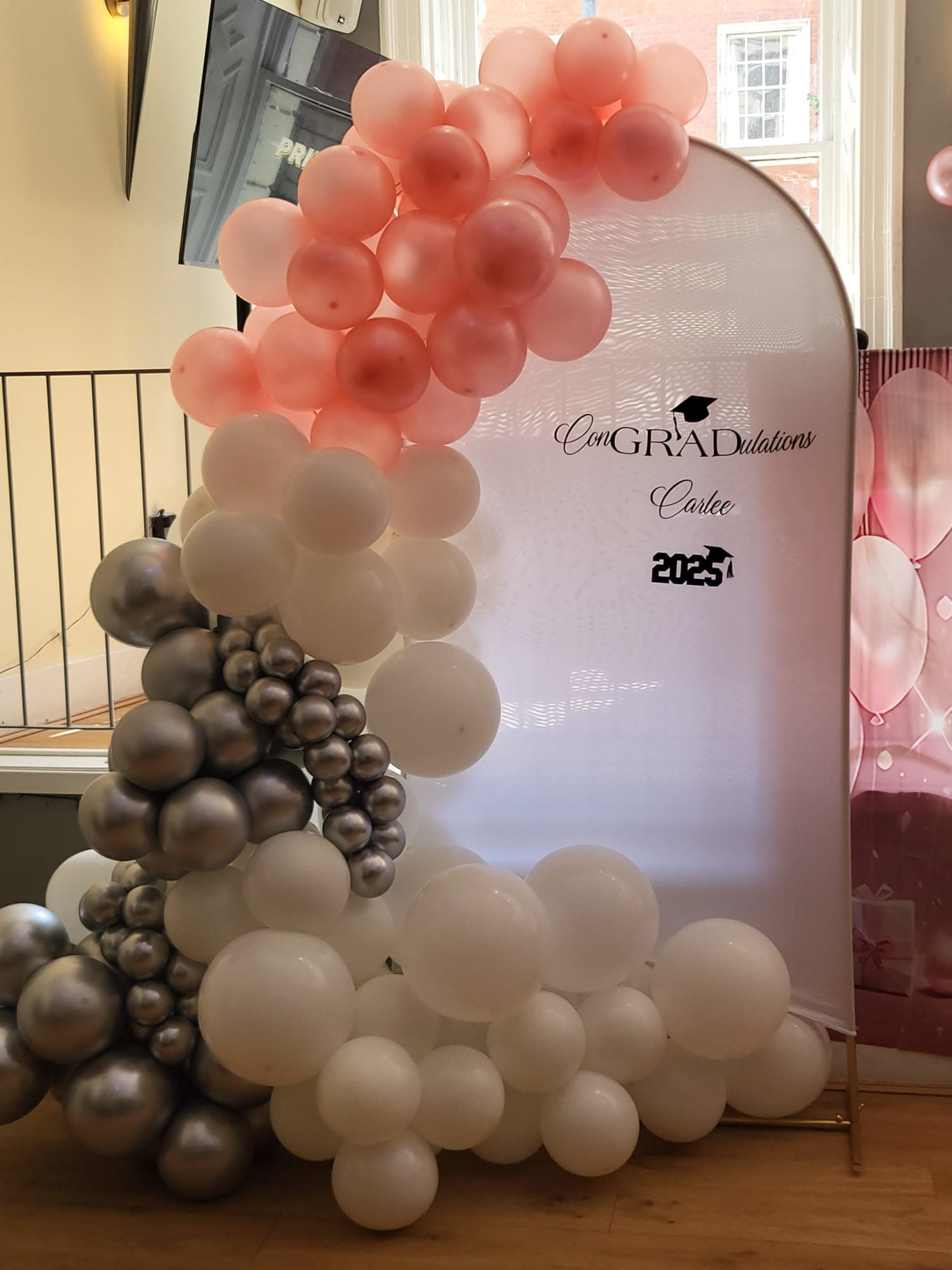 The image shows a decorative arch made of pink, white, and silver balloons, featuring the word "Congratulations" prominently displayed.