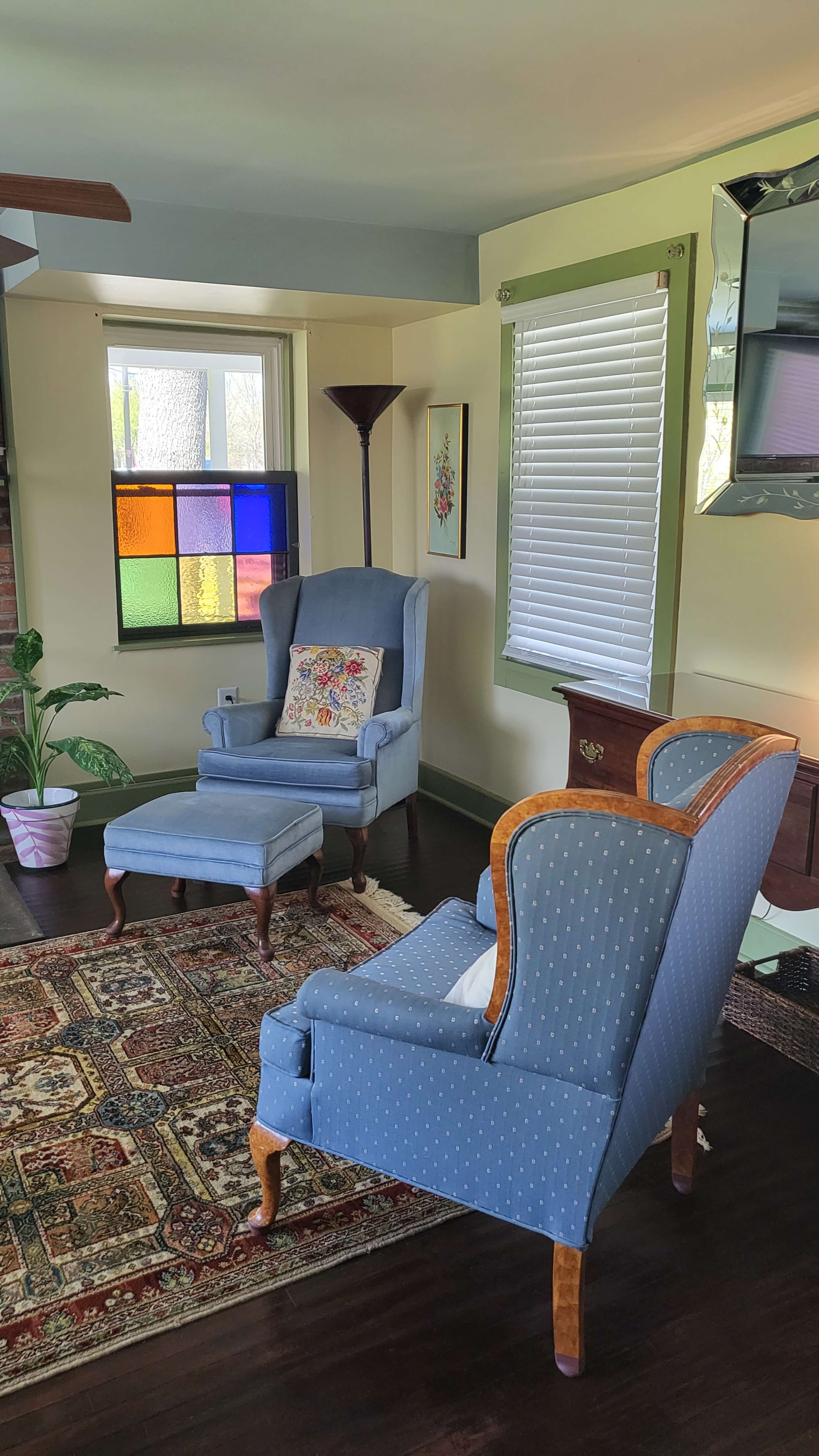 A cozy living room features two blue upholstered chairs, a matching ottoman, a colorful stained glass window, and a patterned area rug.