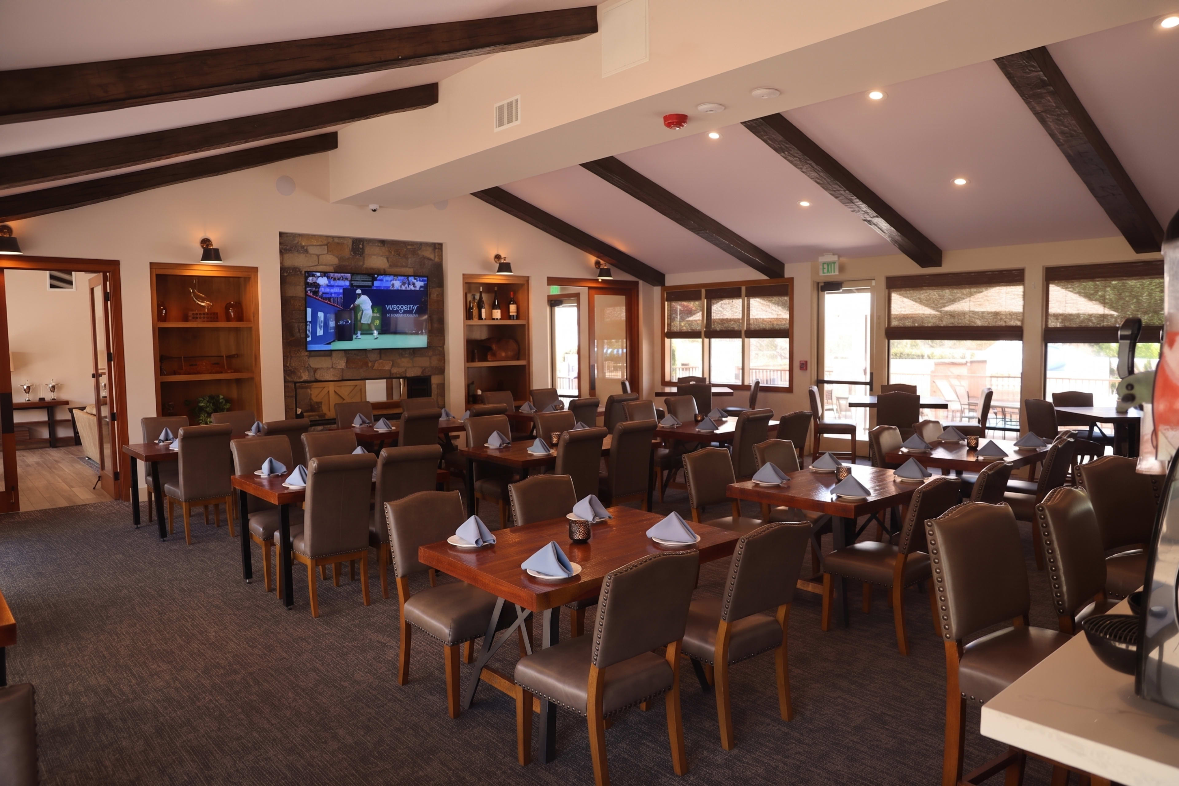 The image shows a spacious dining area with wooden beams, neatly arranged tables, and a television mounted on the wall.