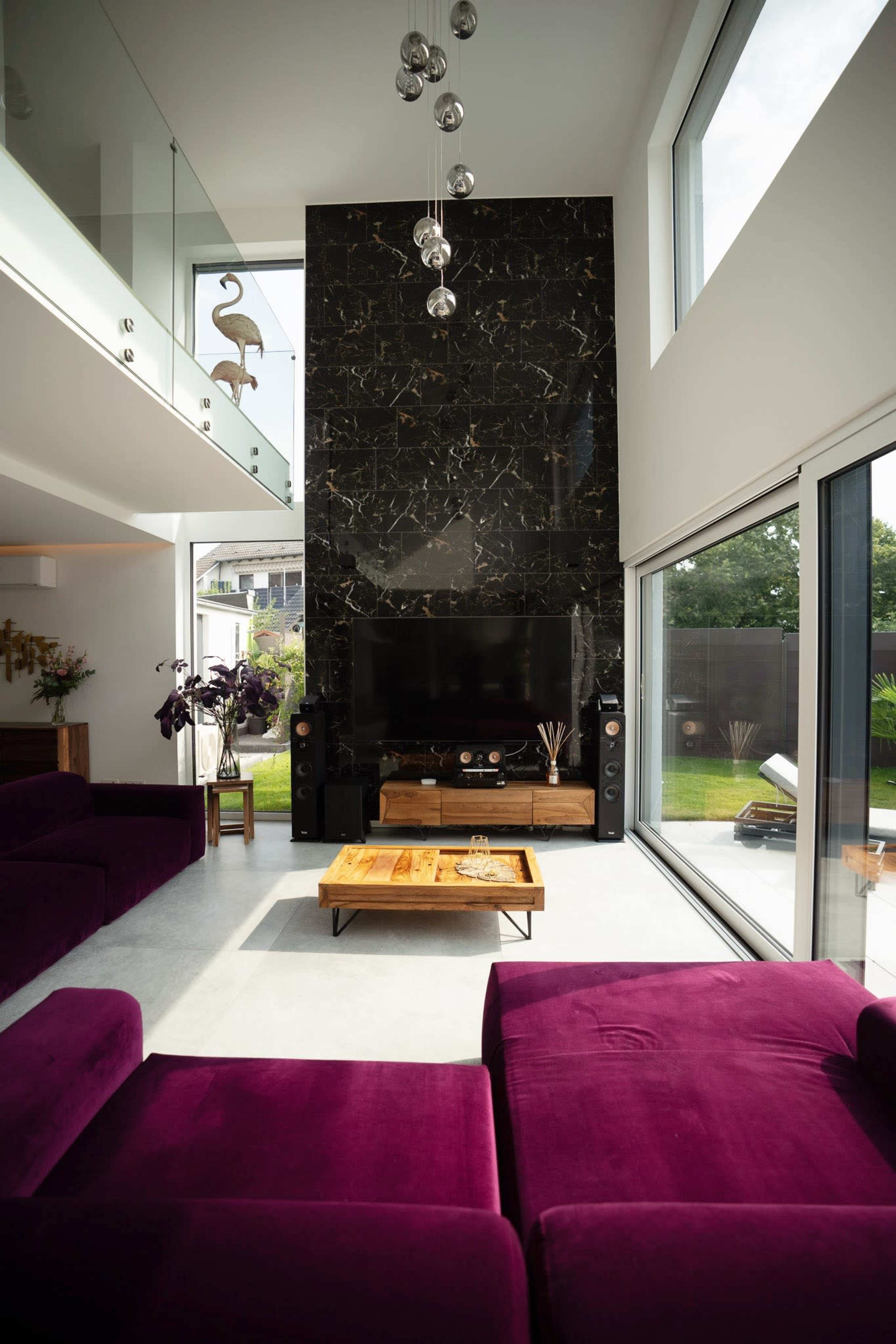 The living room features a tall dark stone wall with a large TV mounted above a wooden media console, surrounded by modern furniture and large windows letting in natural light.
