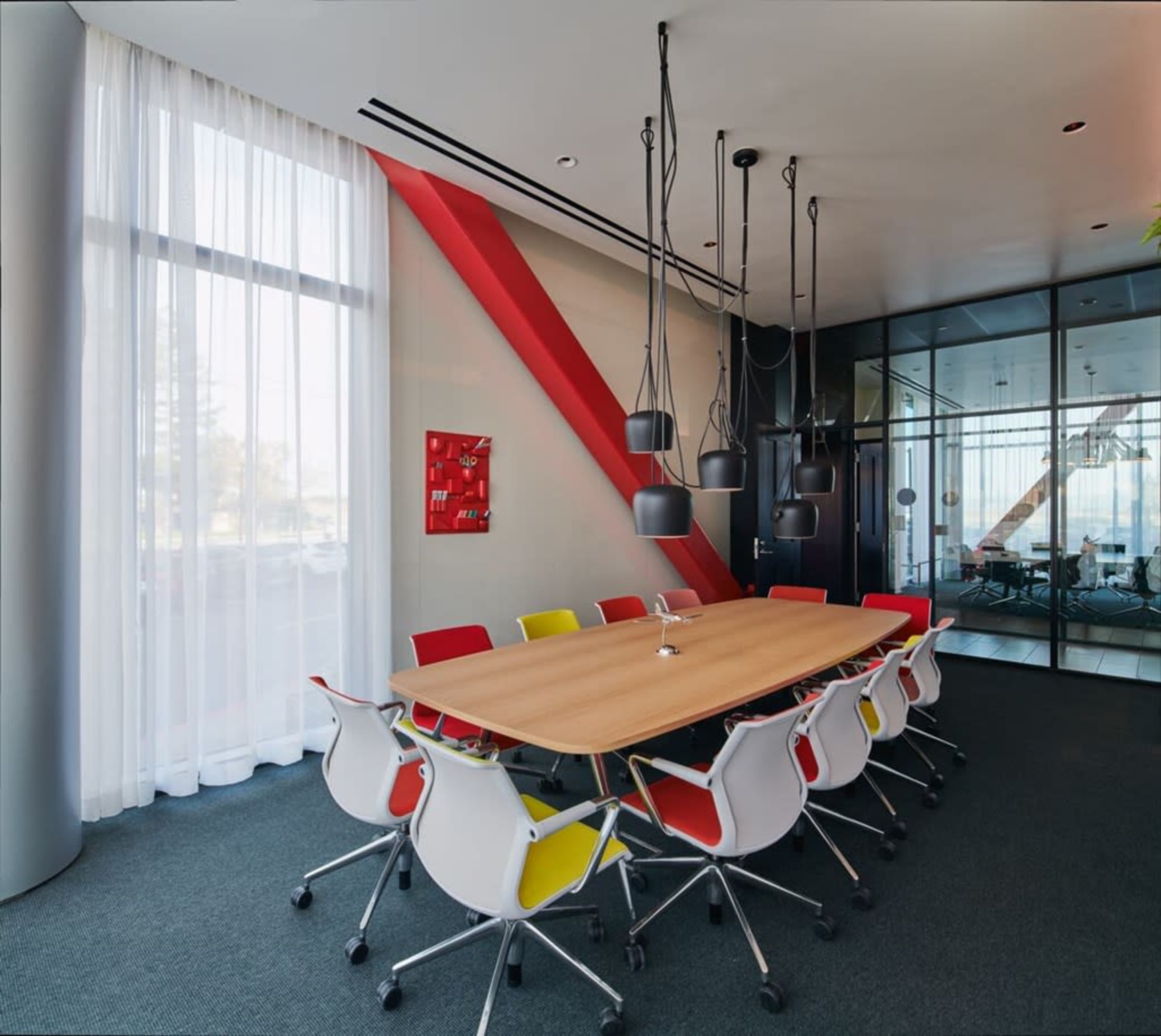 A modern conference room features a large wooden table surrounded by colorful chairs, with large windows allowing natural light to enter.