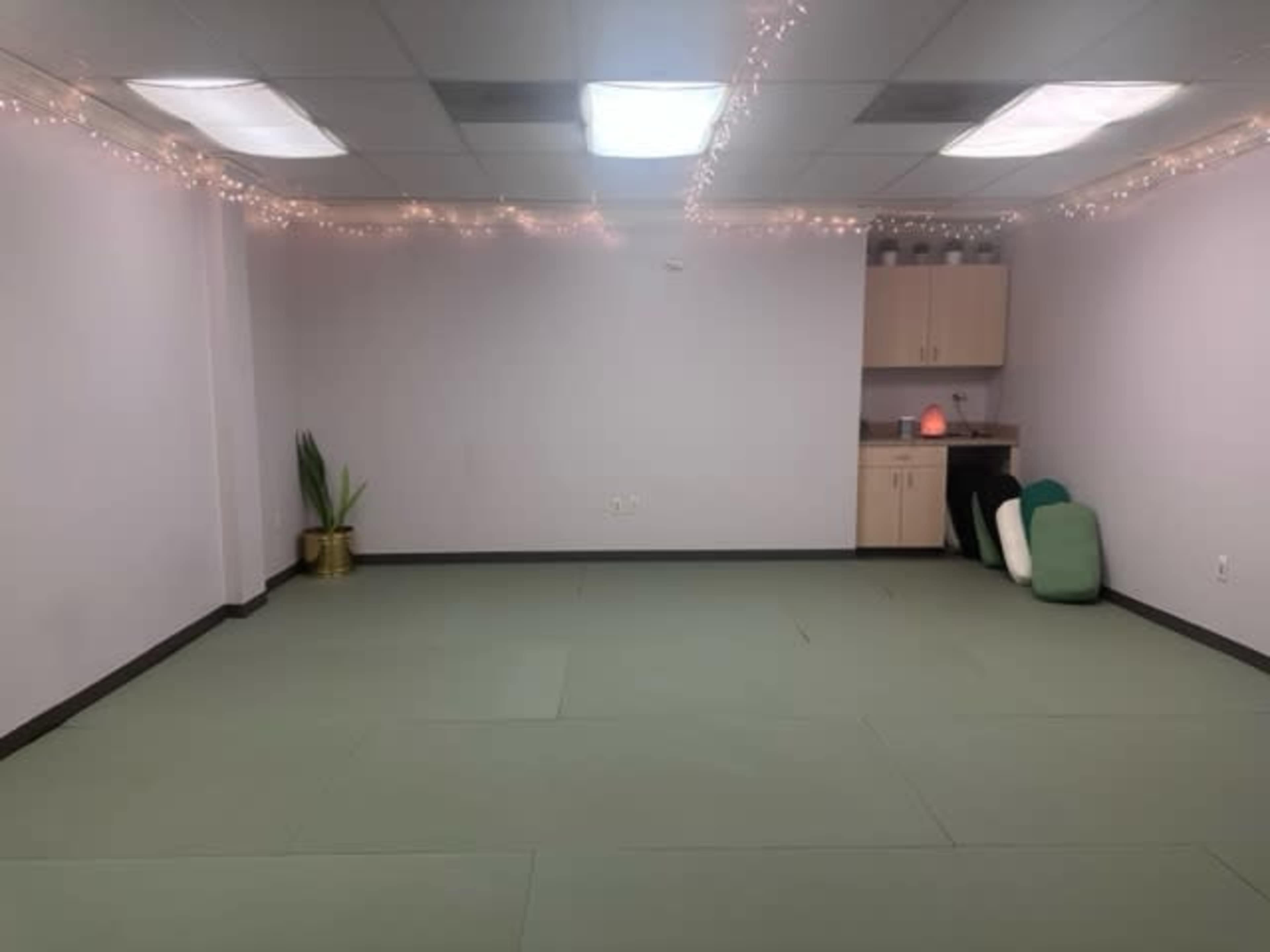 A sparsely furnished room with green mats on the floor, a small kitchen area with cabinets, and string lights along the ceiling.