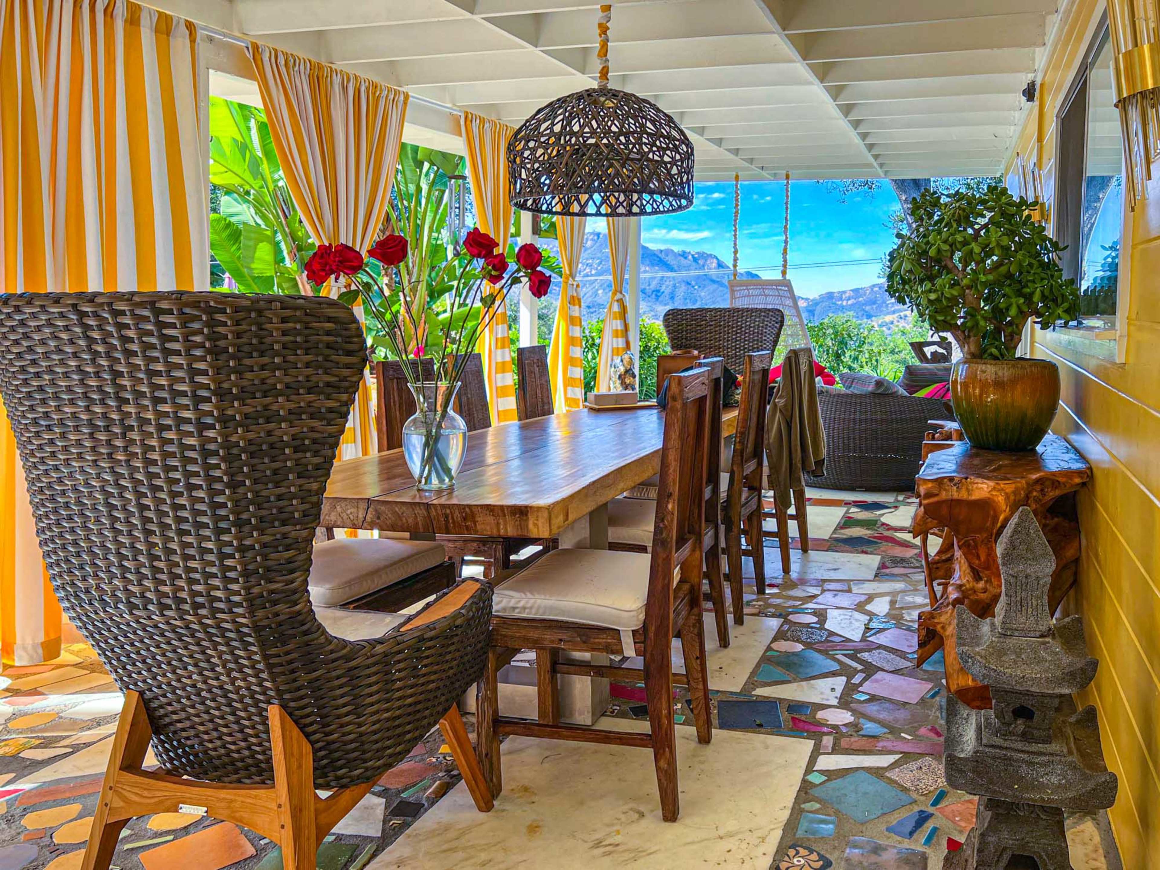 A spacious outdoor dining area features a large wooden table surrounded by wicker chairs, adorned with decorative plants and a view of mountains in the background.