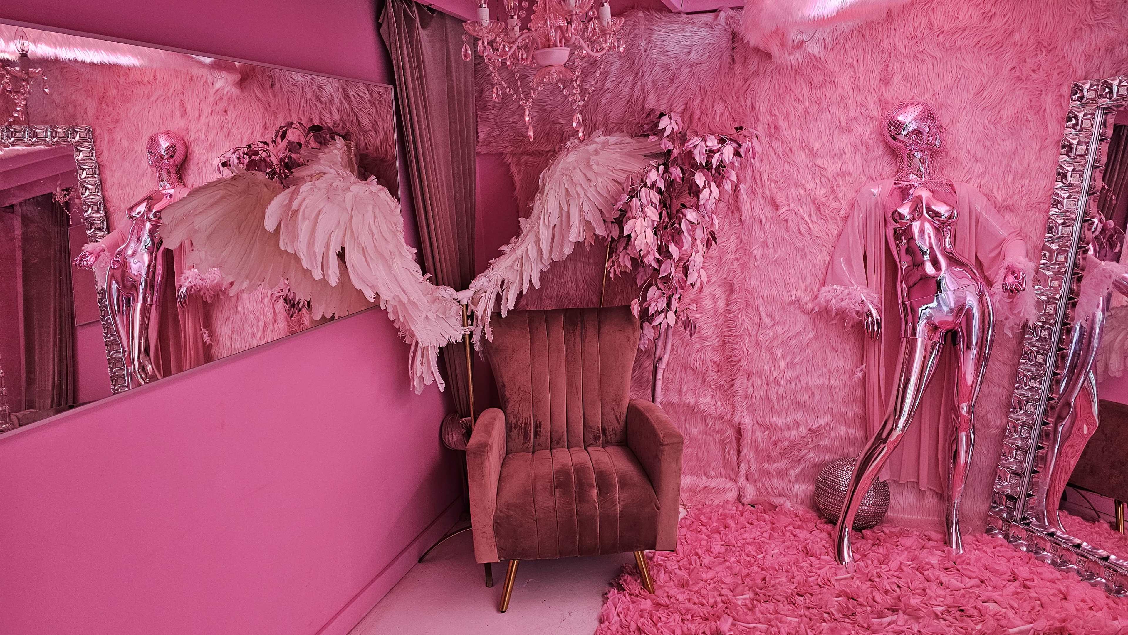 The room features a pink color scheme with a velvet chair, mirrored decorations, a mannequin in shiny attire, and angel wings displayed against a backdrop of fluffy texture.