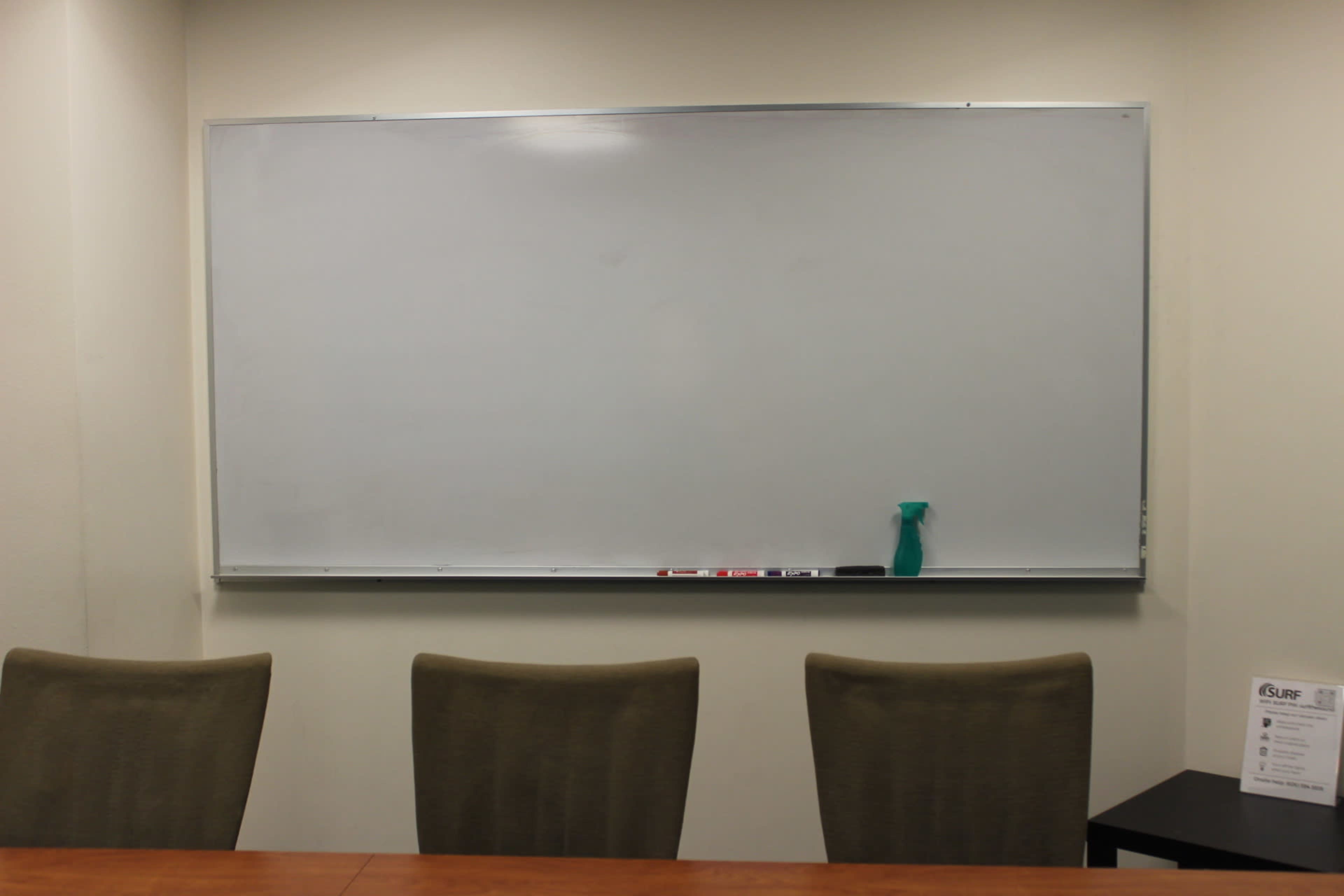 A clean whiteboard is mounted on a wall above a wooden table with three empty chairs in front of it.