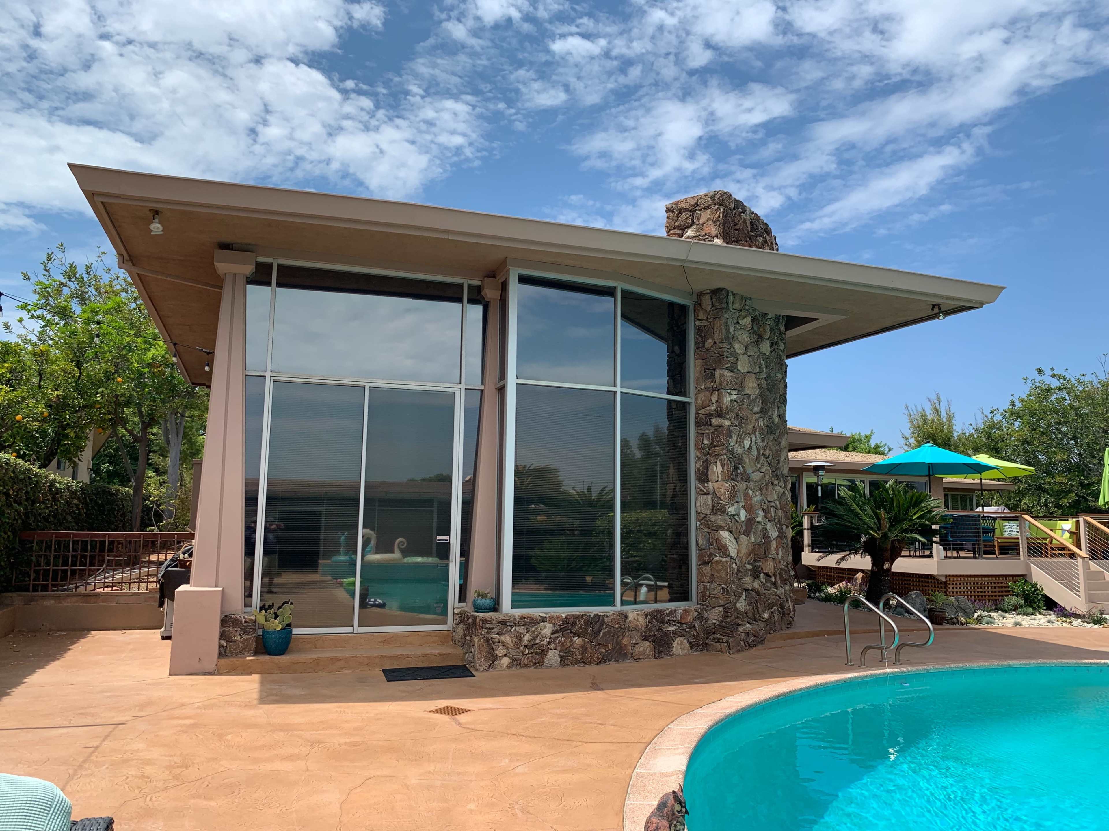 A modern house with large glass windows and a stone chimney is situated beside a swimming pool surrounded by landscaping.