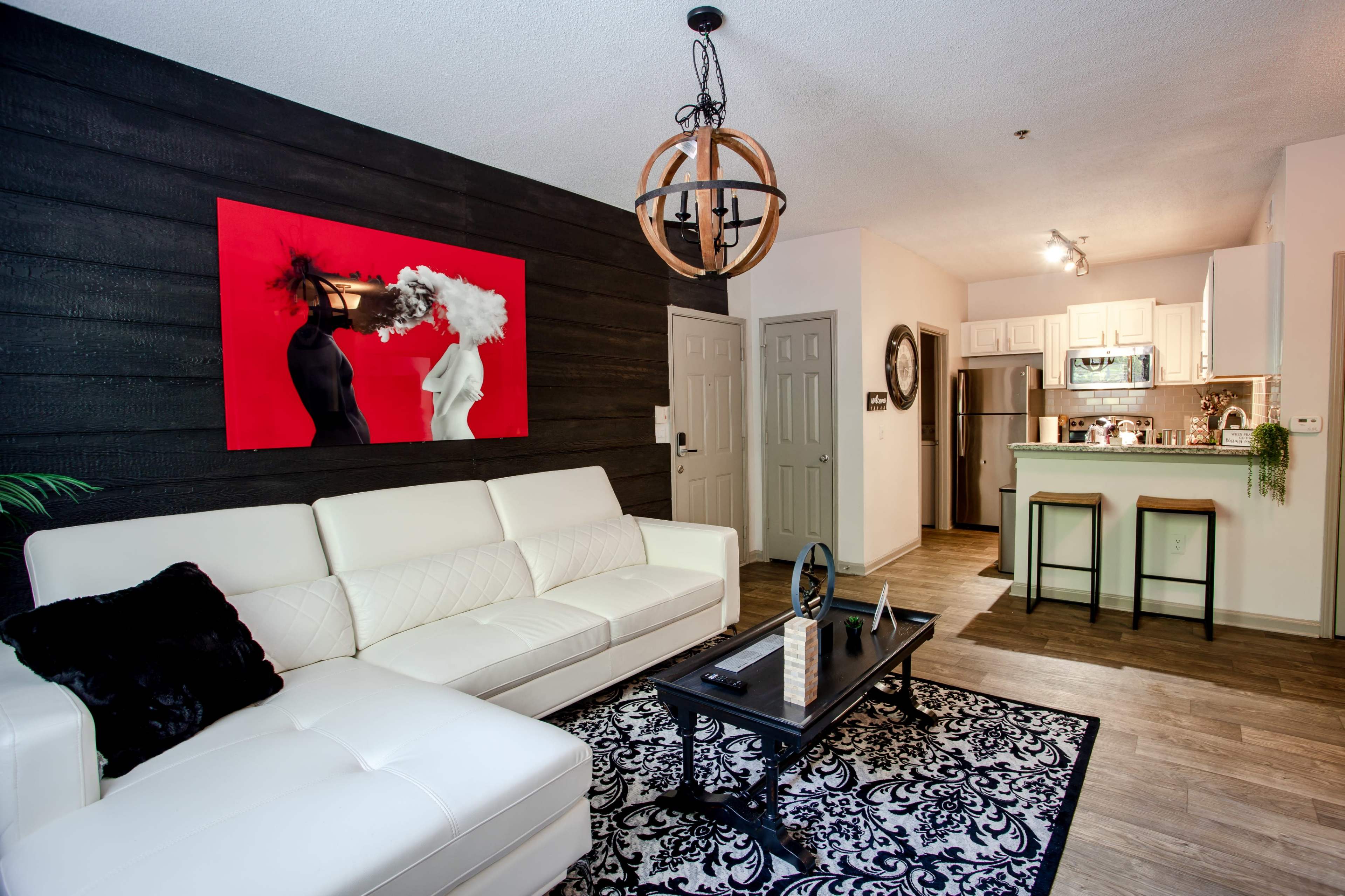 A modern living room with a white sectional sofa, a black coffee table on a patterned rug, and a dining area in the background featuring a round light fixture and a kitchen with white cabinets.