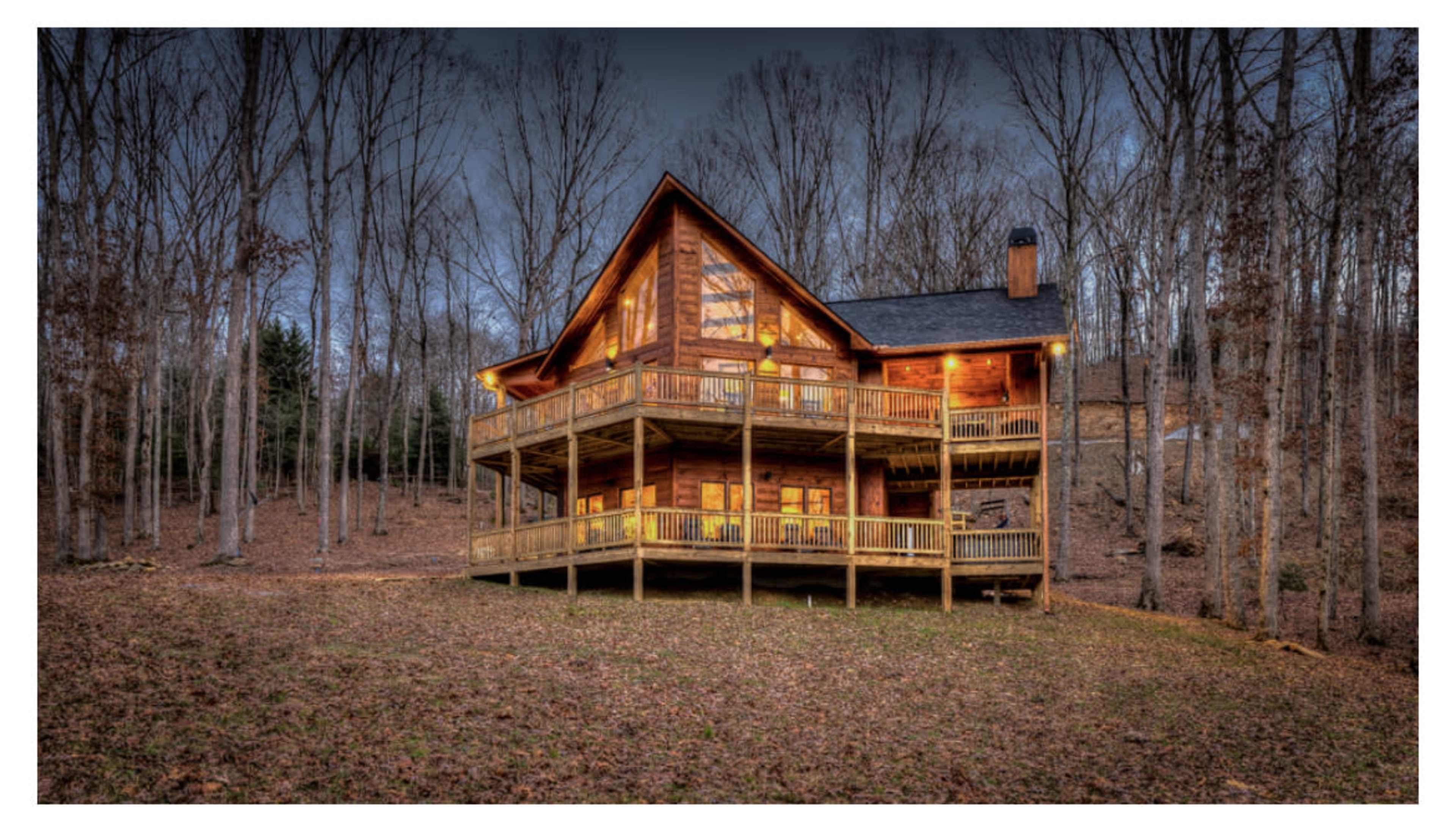 A large wooden cabin with multiple balconies set among bare trees in a wooded area during dusk.