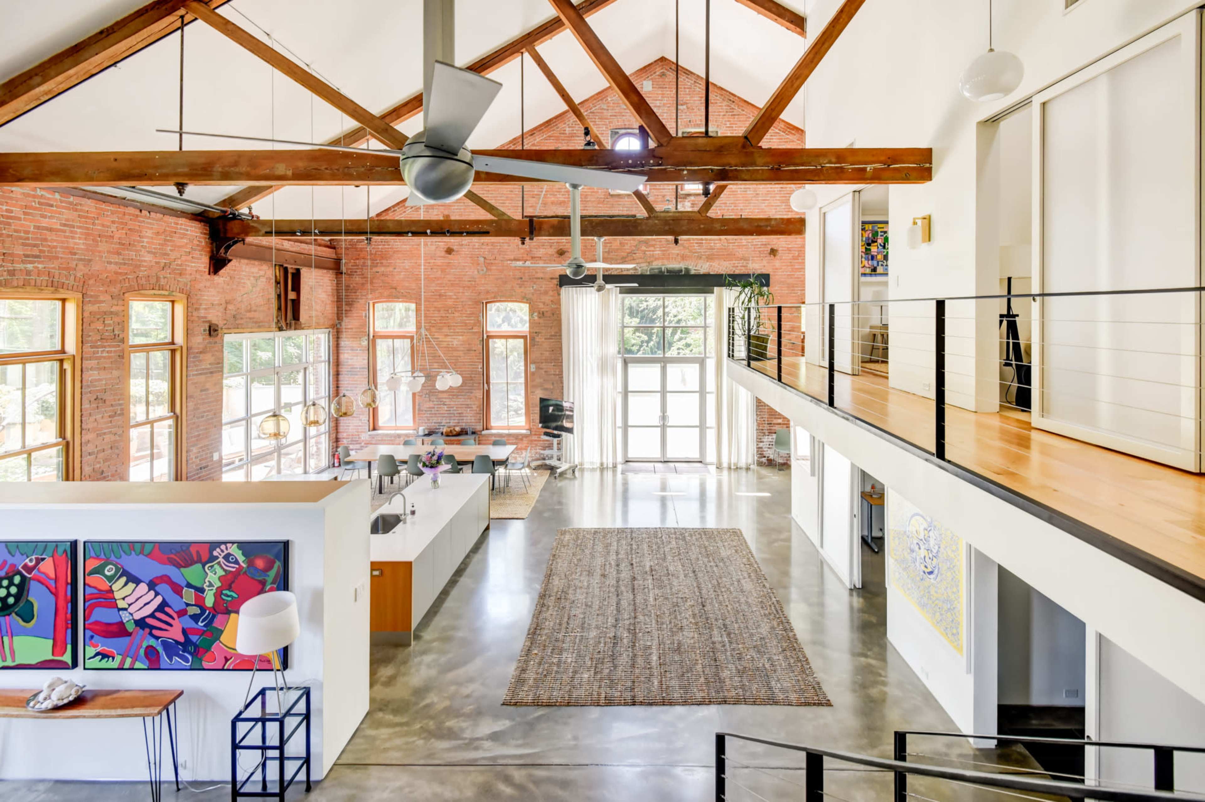 The image shows a spacious, modern interior with high ceilings, exposed brick walls, and a large open area featuring a kitchen, living space, and a balcony overlooking the first floor.