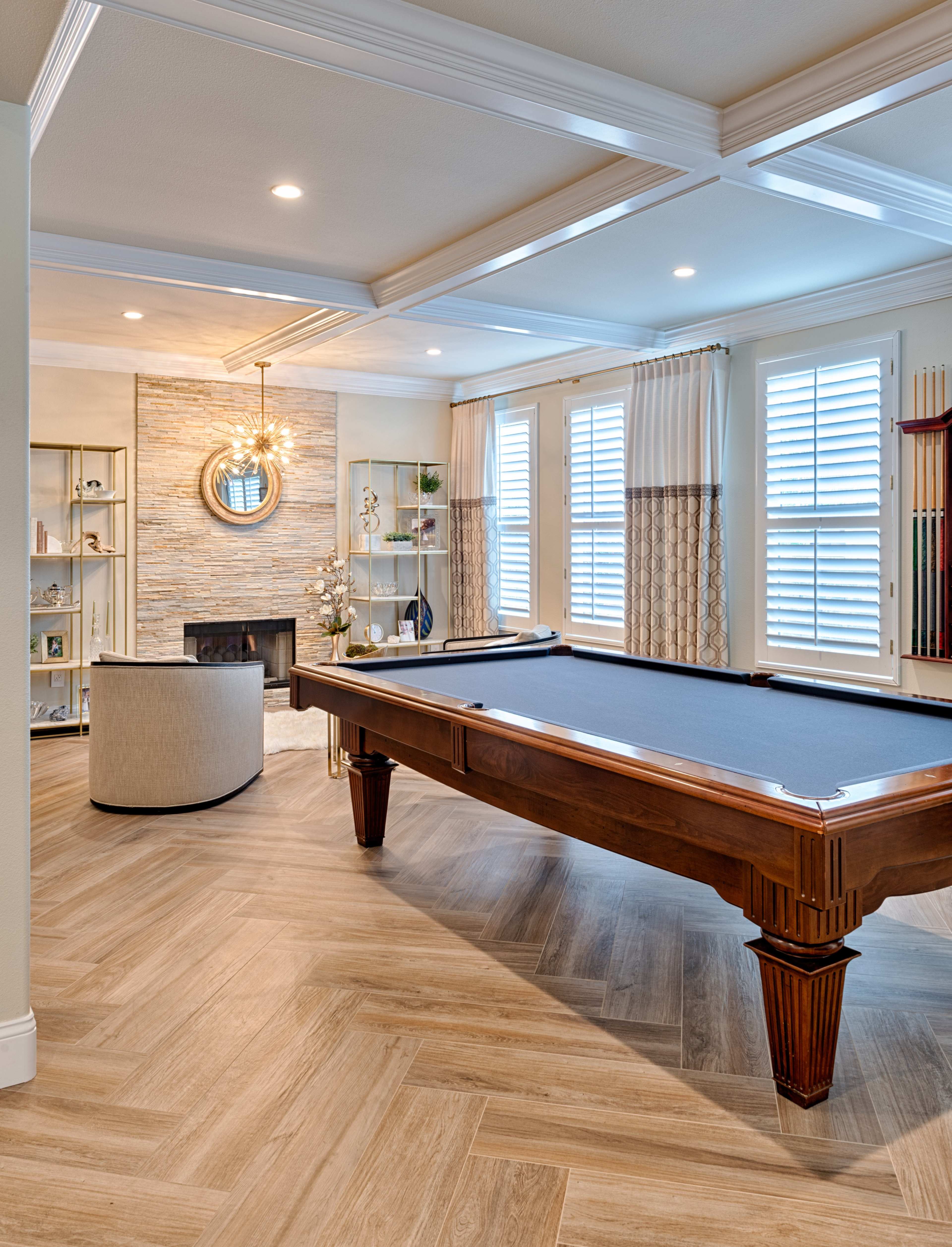 The image shows a modern game room featuring a billiards table, a cozy seating area, and a stone fireplace surrounded by large windows.