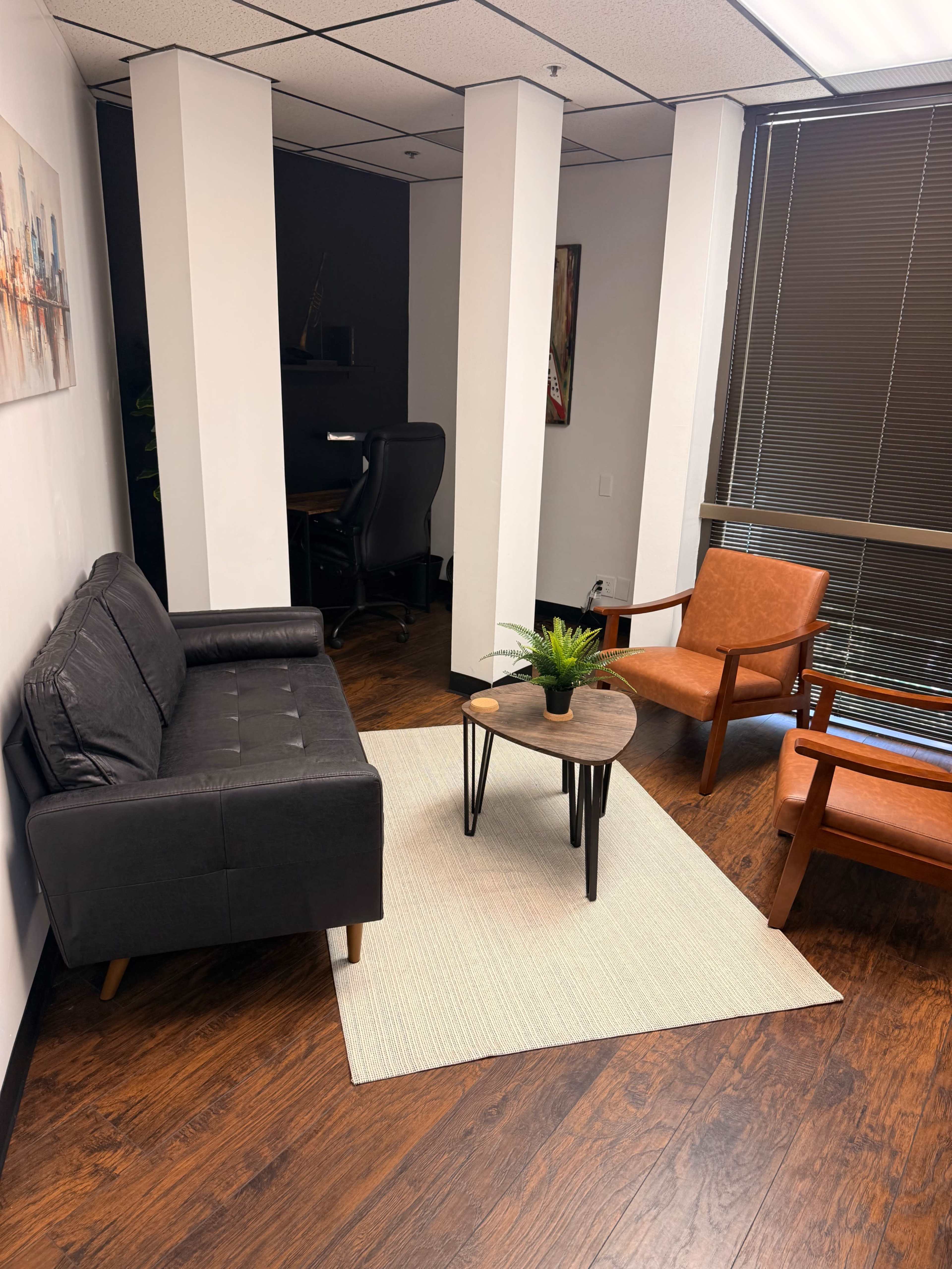The image shows a waiting area with a dark sofa, two brown chairs, a small table with a plant, and a rug on a wooden floor, along with a desk and chair in the background.