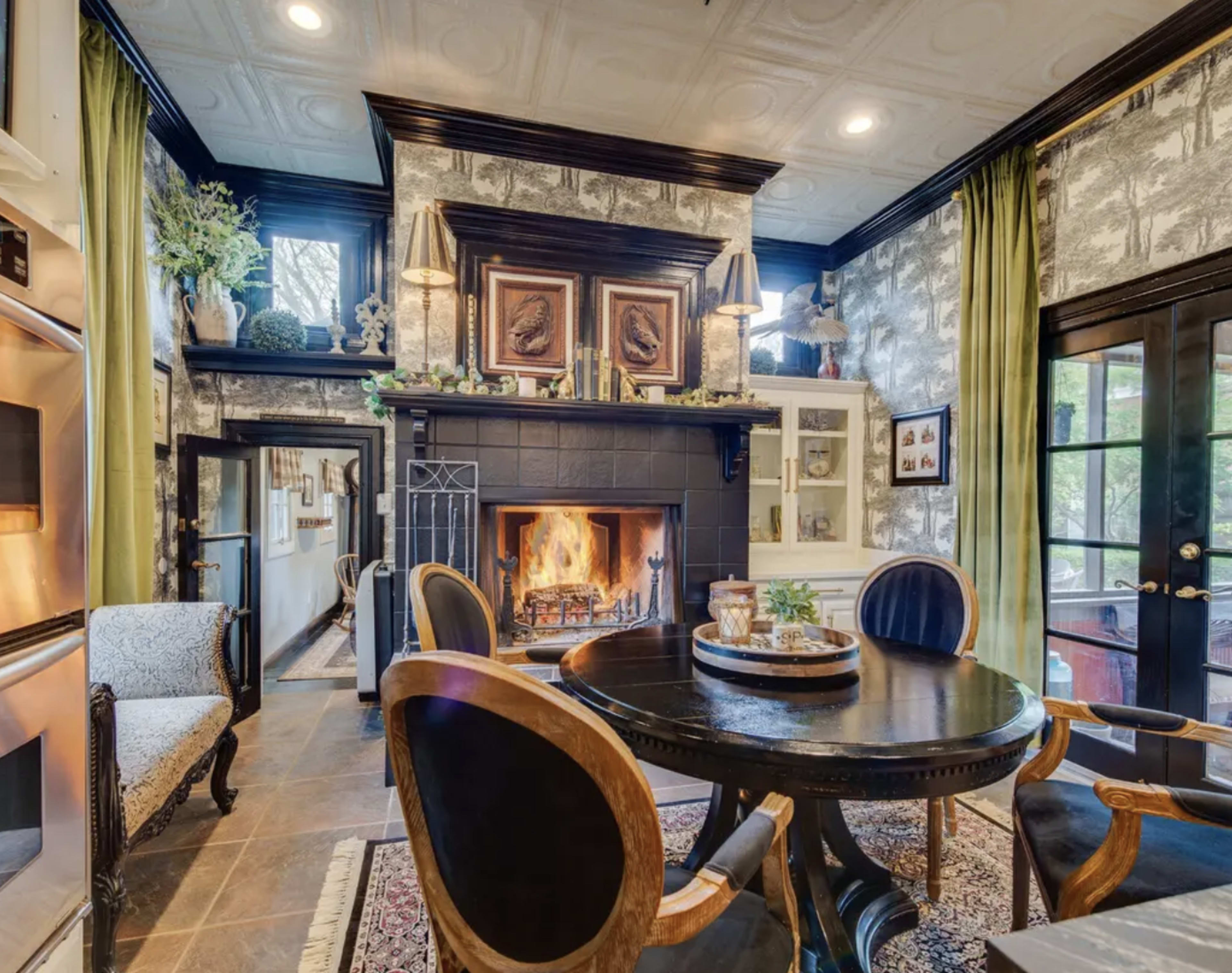 A dining area with a round table and black chairs is positioned near a fireplace, surrounded by patterned wallpaper and green curtains.