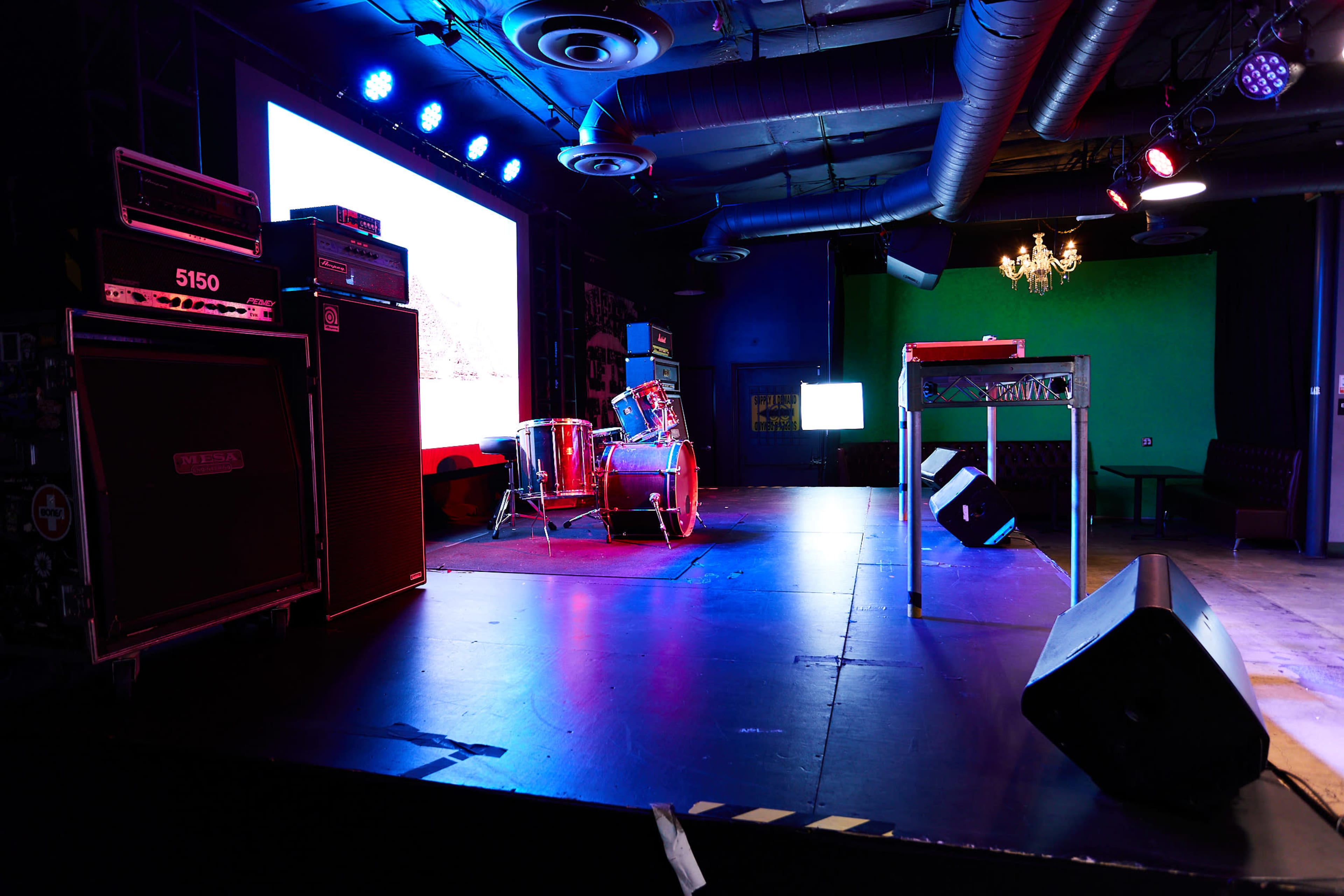 An empty stage set up with musical equipment, including amplifiers, a drum kit, and screens displaying visuals, in a dimly lit venue.