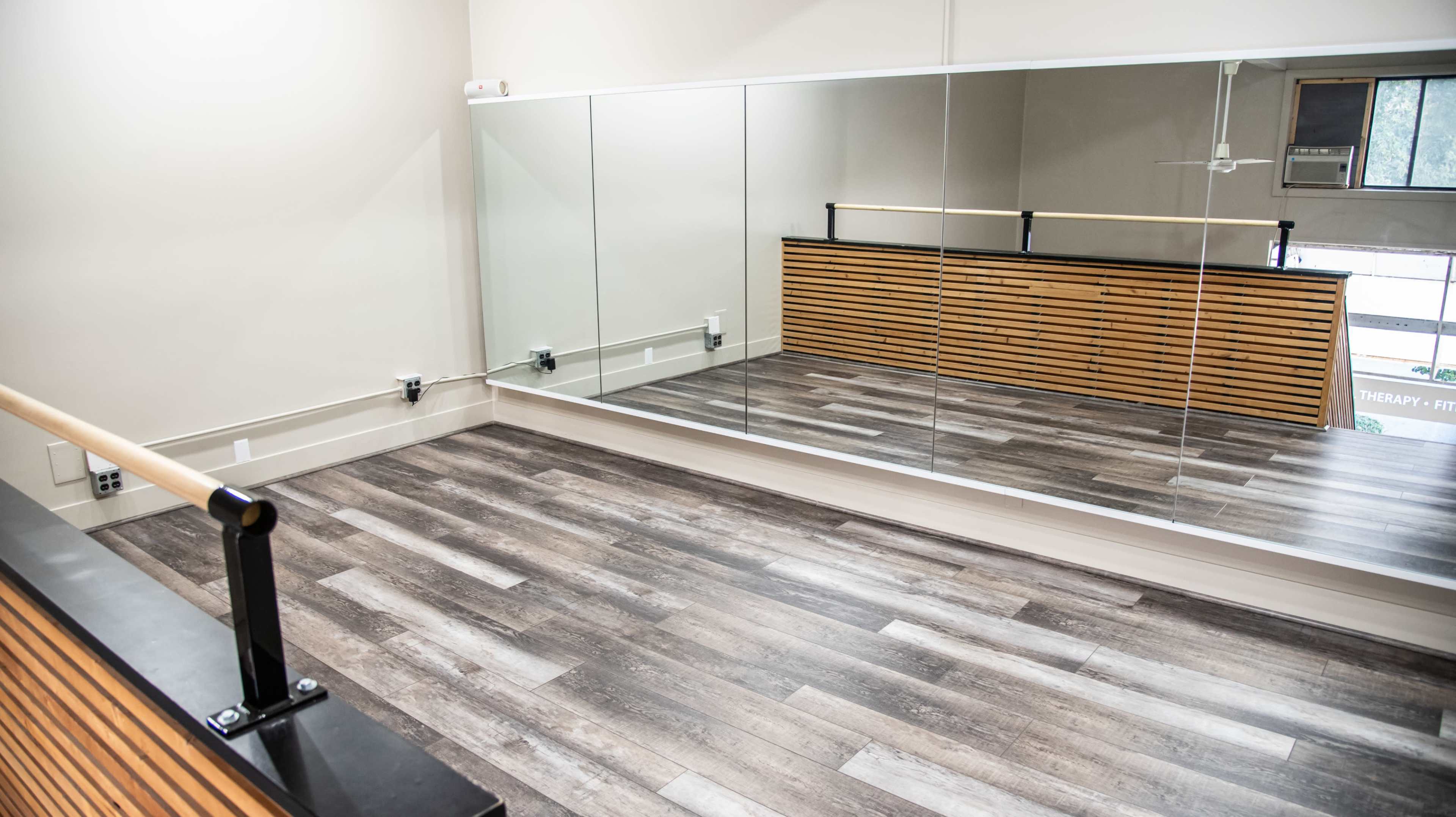 A minimalist therapy or dance studio featuring a mirrored wall, wooden accents, and a spacious wooden floor.