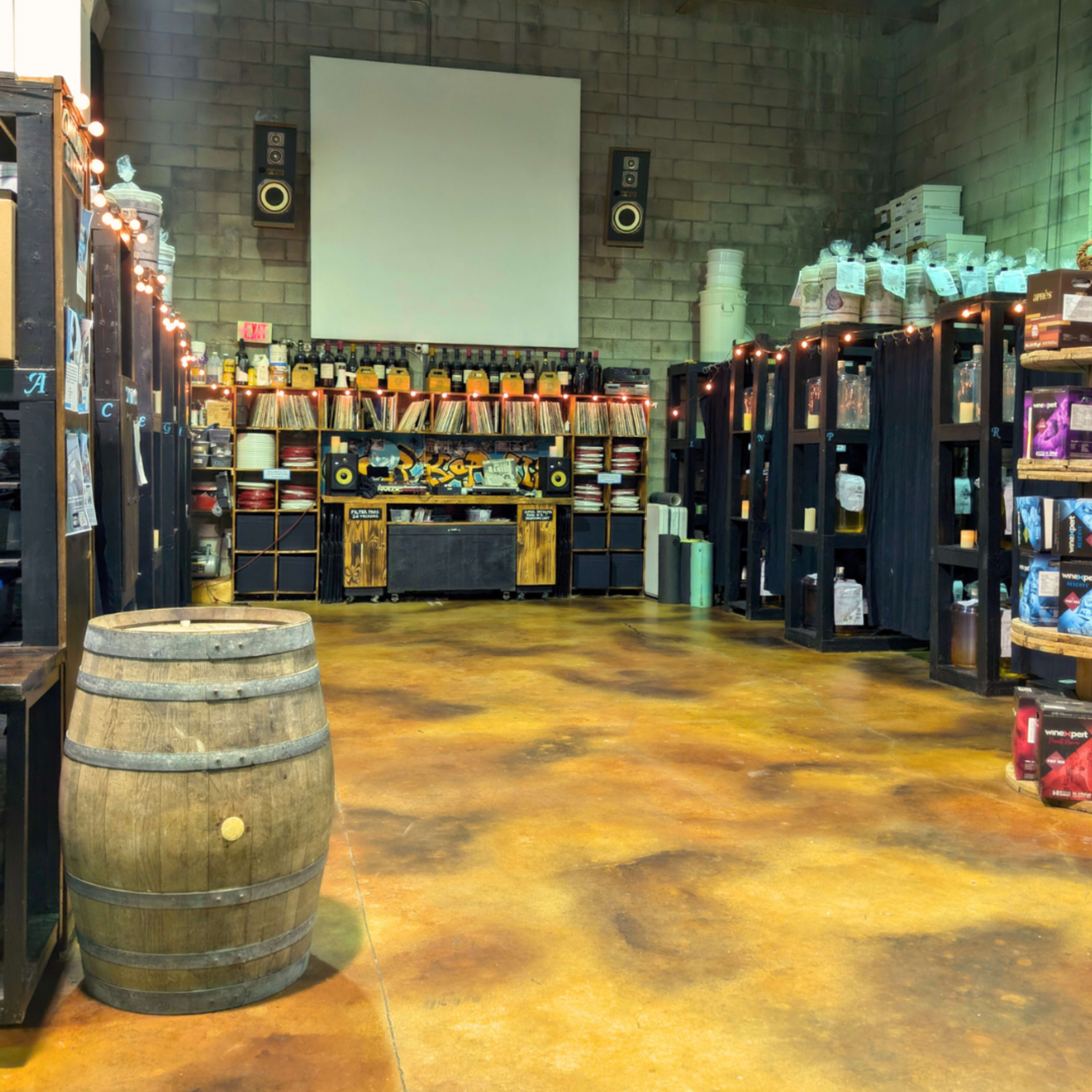 The image shows a spacious interior of a warehouse with shelves filled with various containers and a wooden barrel placed in the foreground.