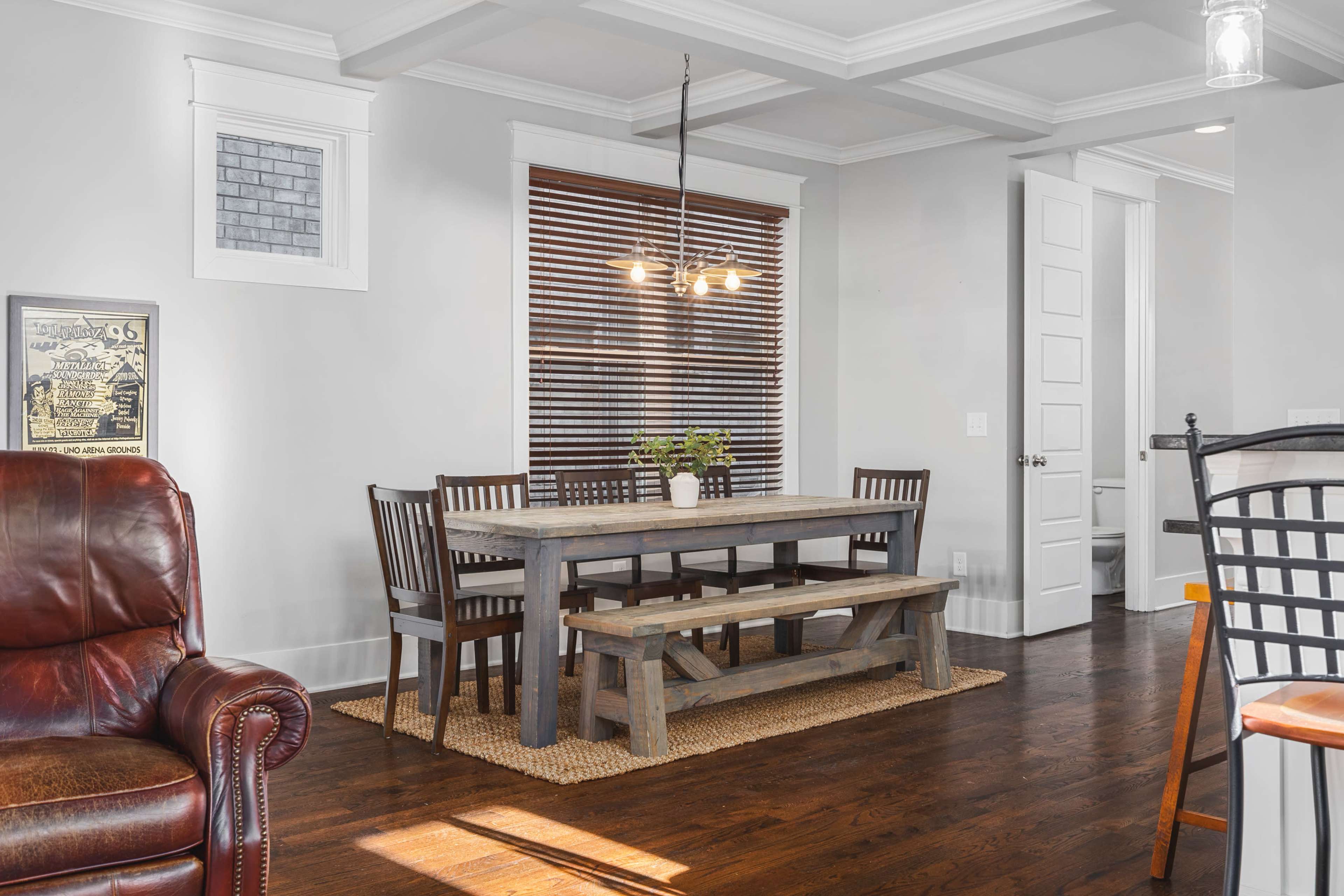 A wooden dining table with benches is set in a well-lit room featuring a window with blinds, brown hardwood flooring, and a leather armchair in the foreground.