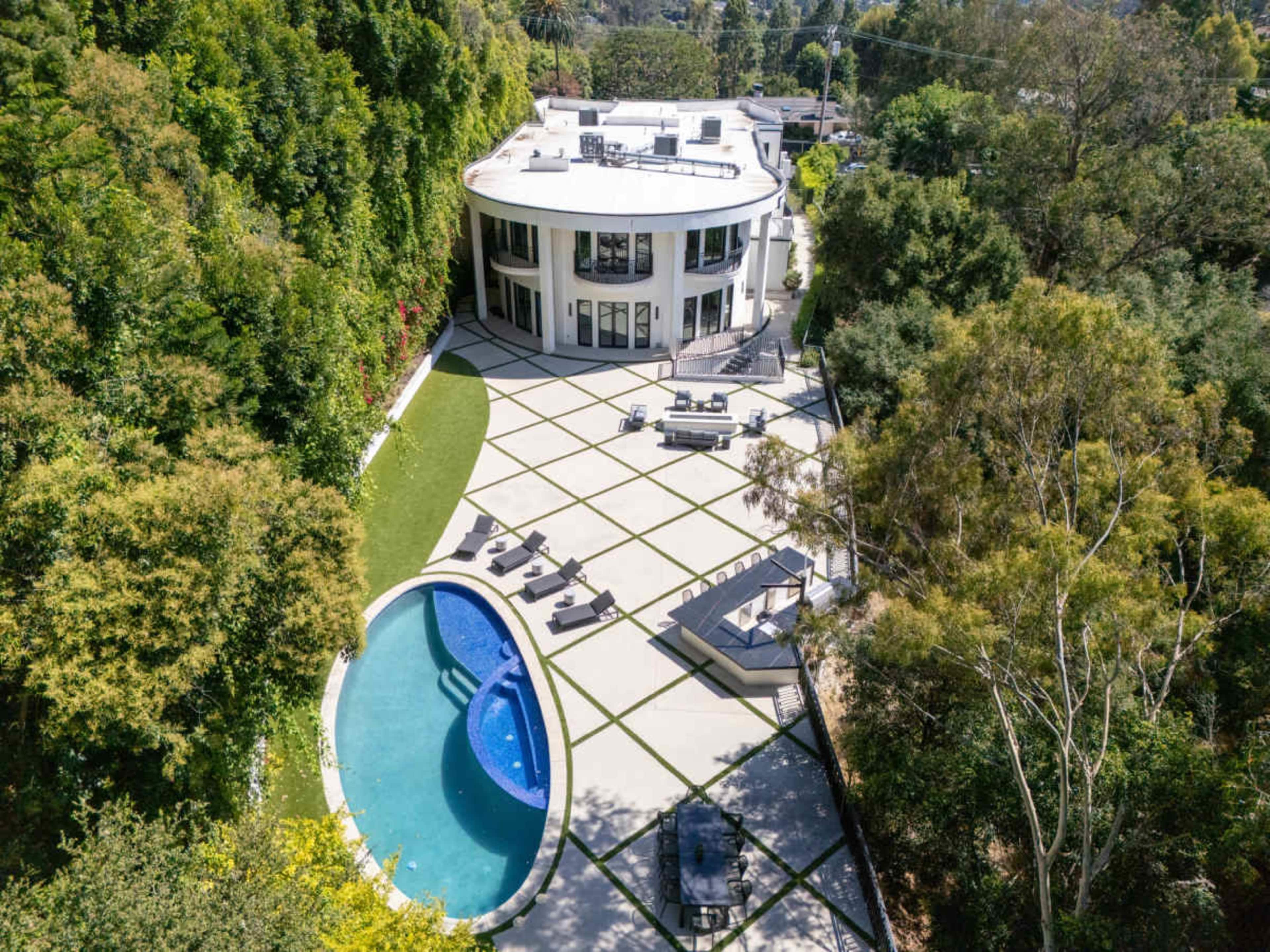 The image shows a modern, circular house surrounded by greenery, featuring a pool and outdoor seating areas arranged on a geometric patio.