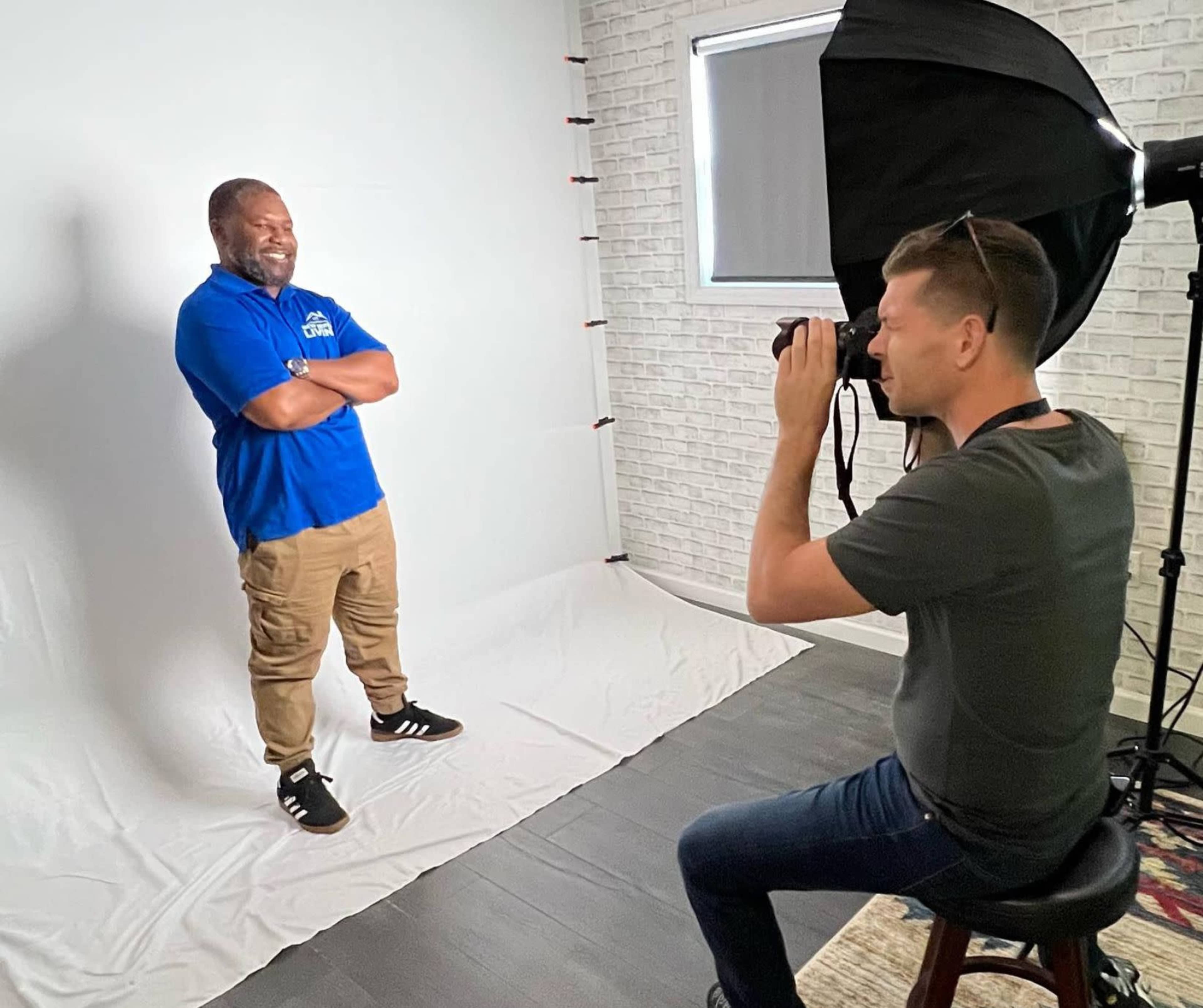 A photographer captures a smiling man in a blue shirt while he poses with his arms crossed in a studio setting with a white backdrop.