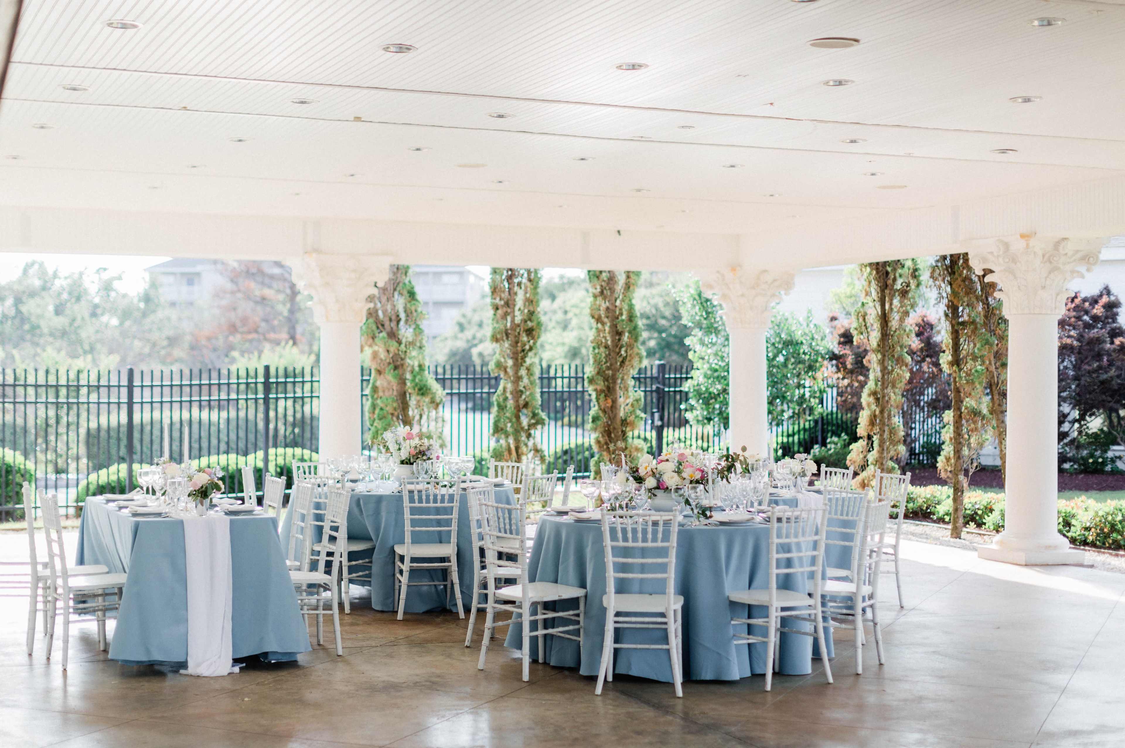 The scene shows a beautifully set outdoor dining area with round tables covered in blue tablecloths, white chairs, and floral centerpieces under a shaded pergola.