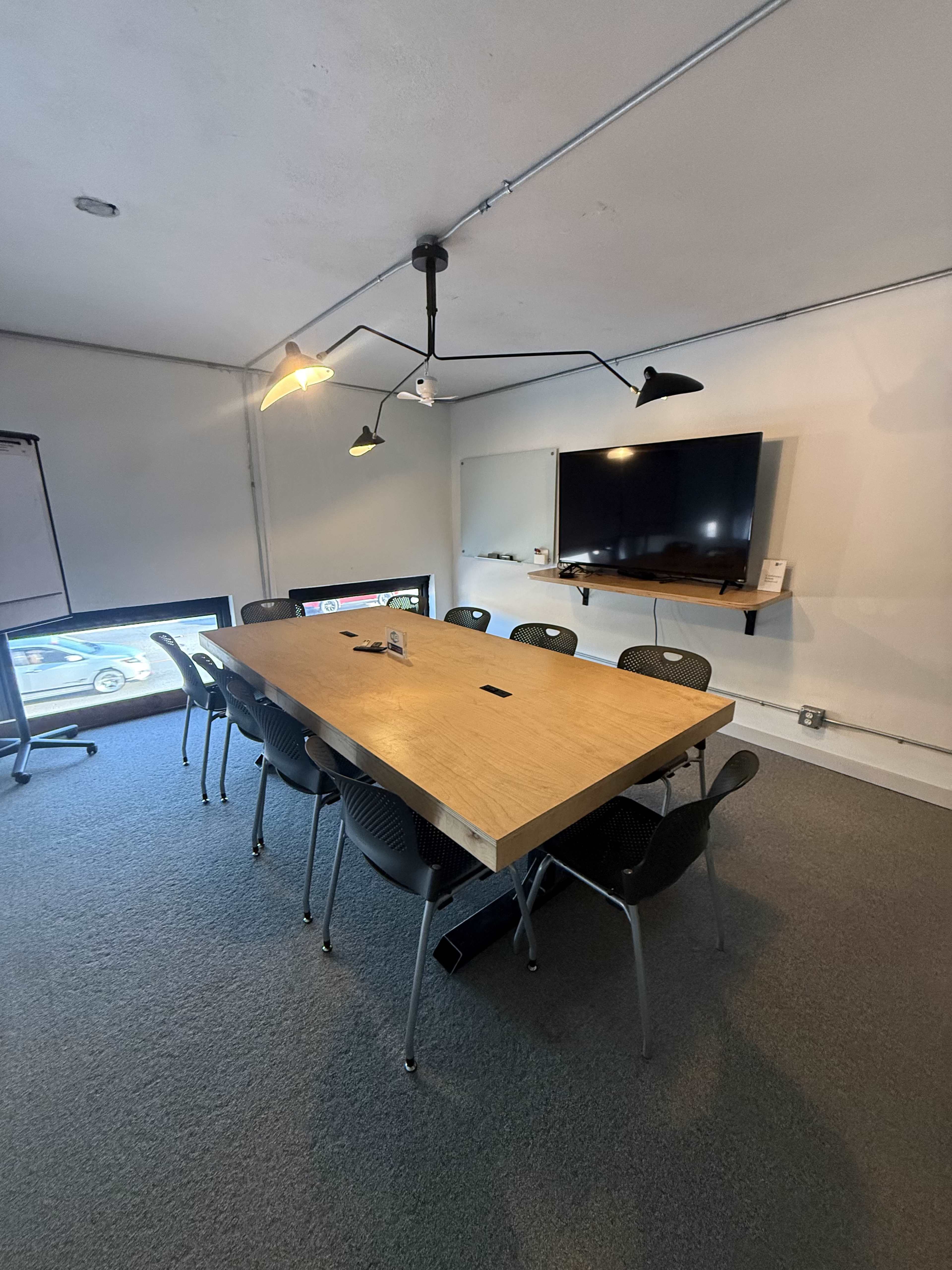 The image shows a conference room with a long wooden table surrounded by black chairs, a TV mounted on the wall, and lamps providing overhead light.