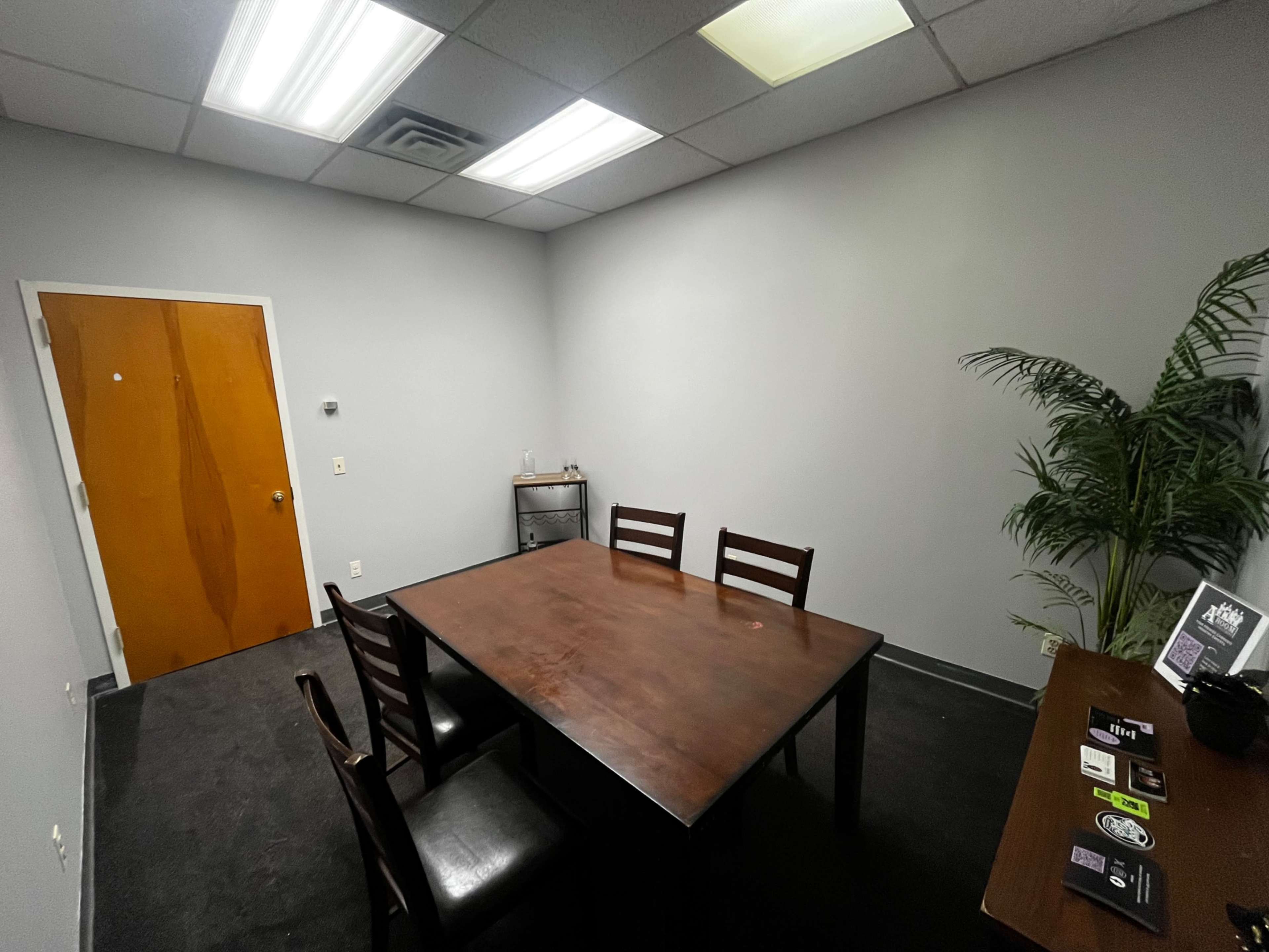 A small, sparsely furnished meeting room with a table and four chairs, a door, a potted plant, and a side table displaying business cards.
