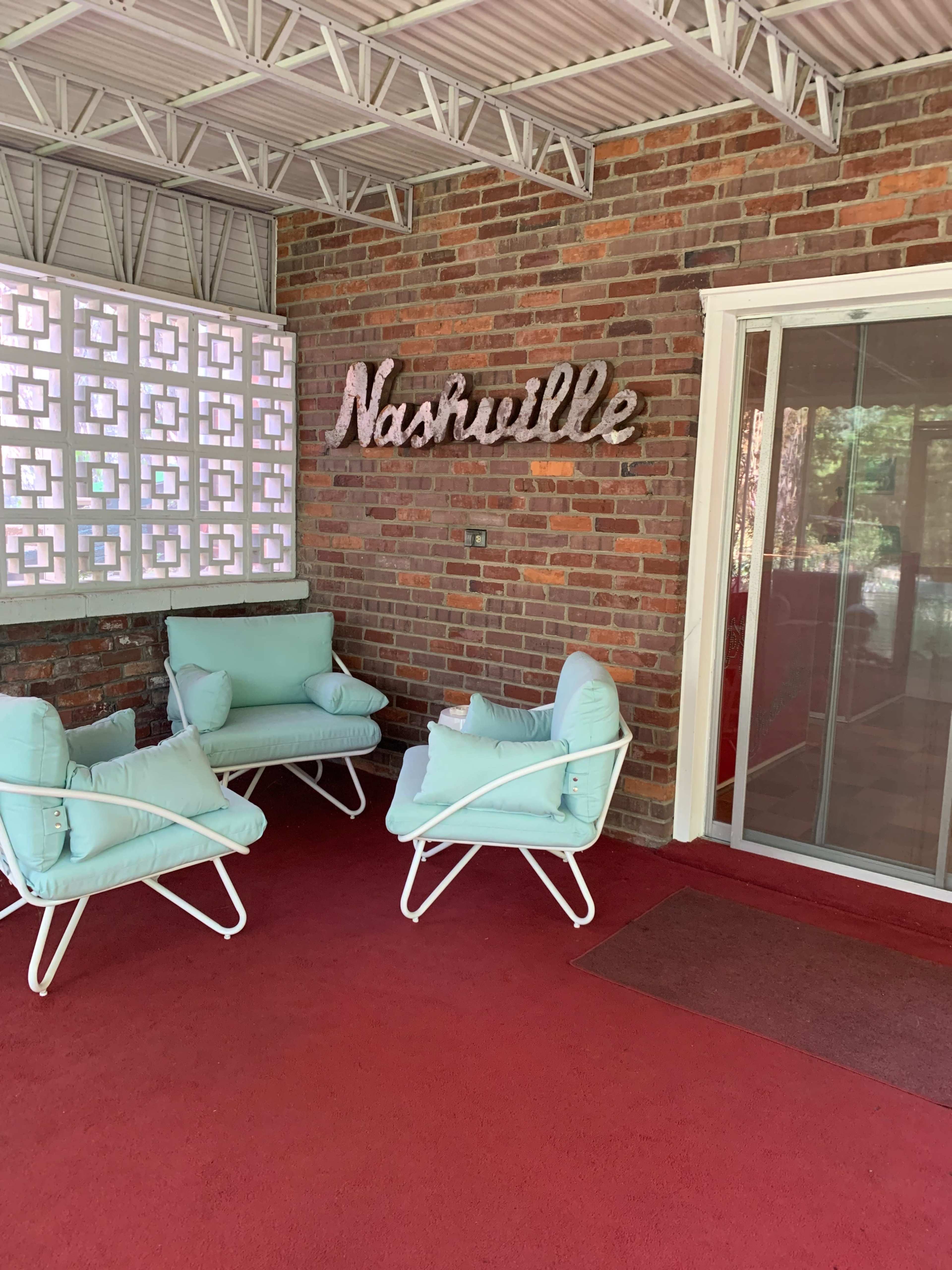 A covered patio features three light blue chairs arranged around a red carpet, with "Nashville" sign displayed on a brick wall.