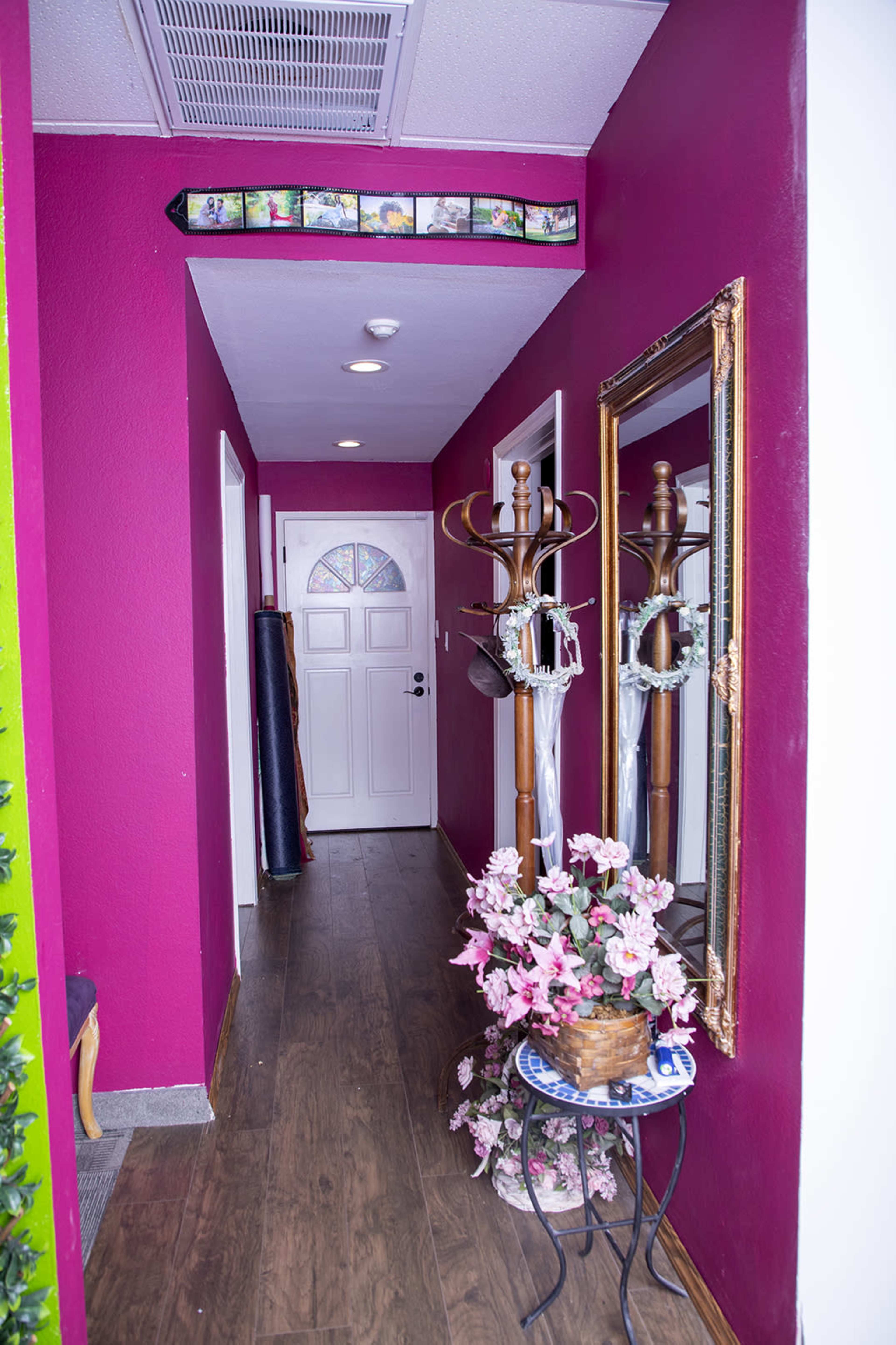 A hallway with a bright pink wall, a front door at the end, a mirror, a coat rack, and a table with flowers.