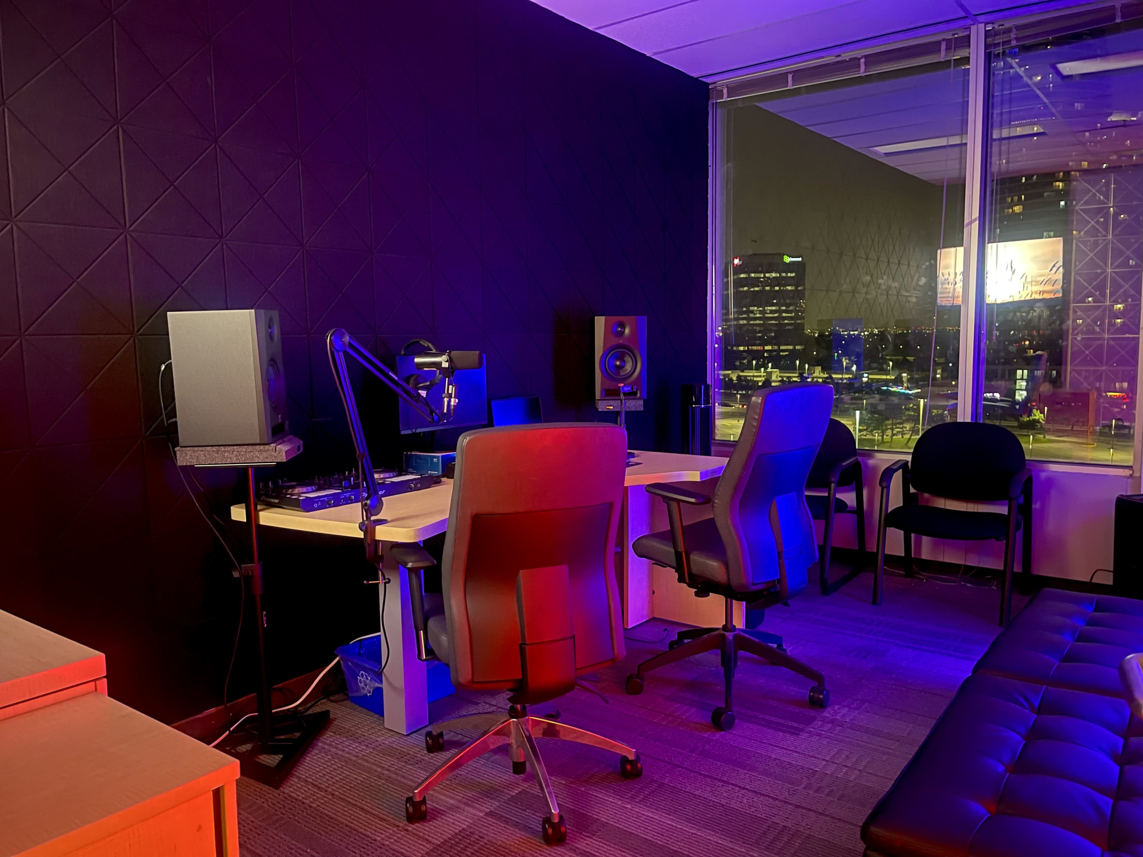 The image shows a dimly lit recording studio with a large desk, two office chairs, speakers, and a view of a city skyline at night through a window.