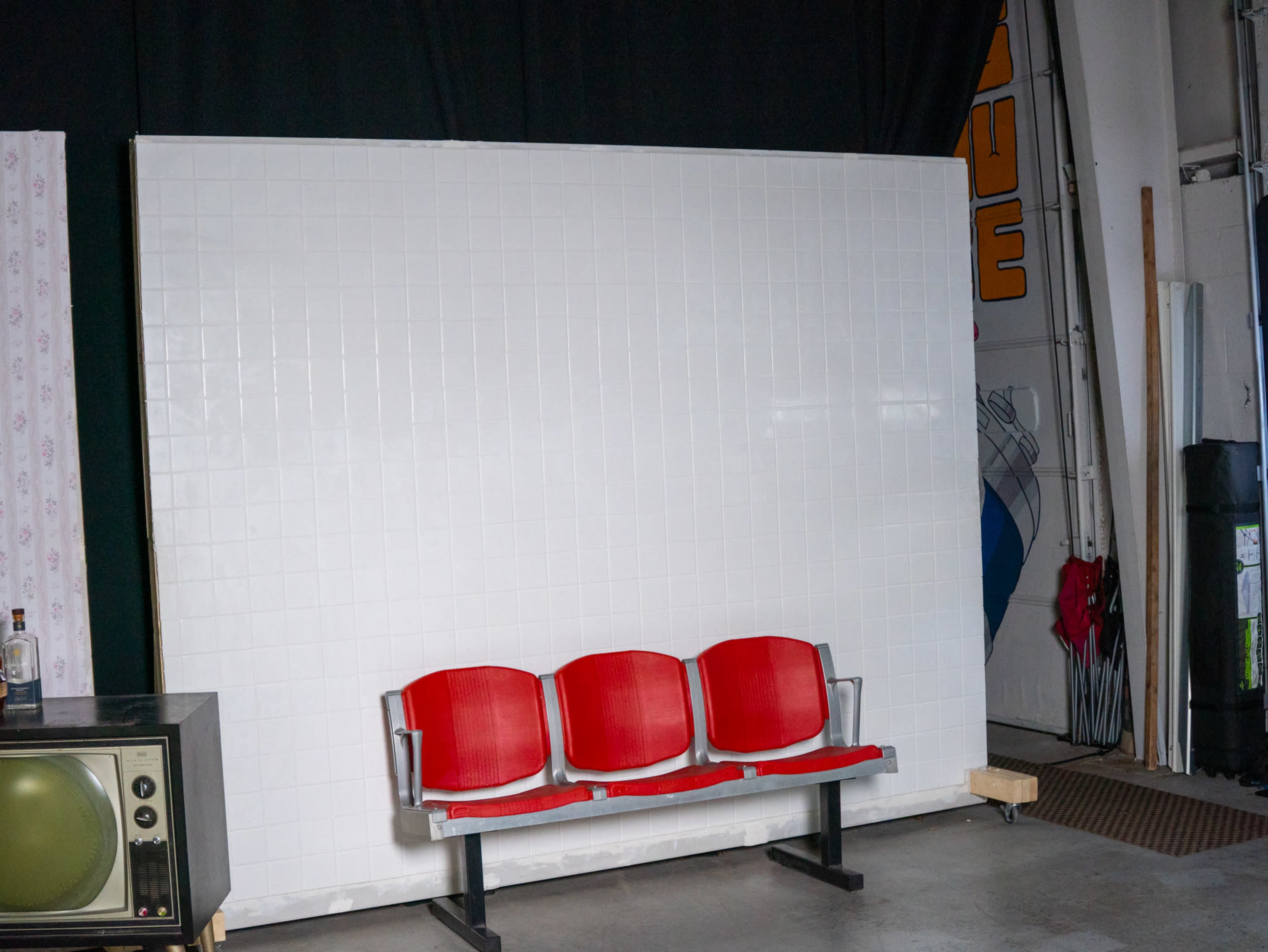The image shows a row of three red chairs in front of a large white wall, with a vintage television set beside them.