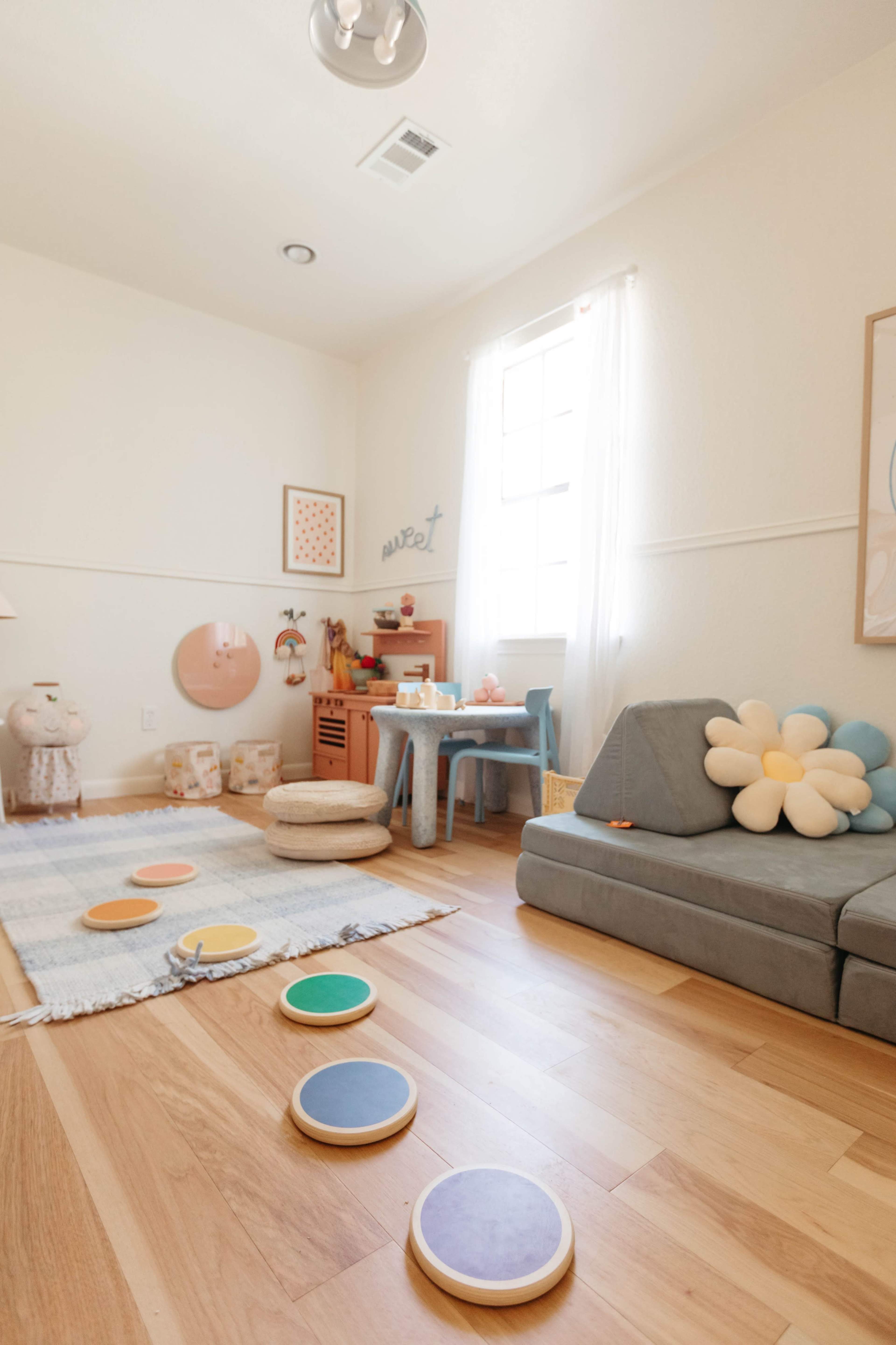 A light and airy playroom with a gray sofa, a blue table, decorative elements, and colorful round mats arranged on the wooden floor.