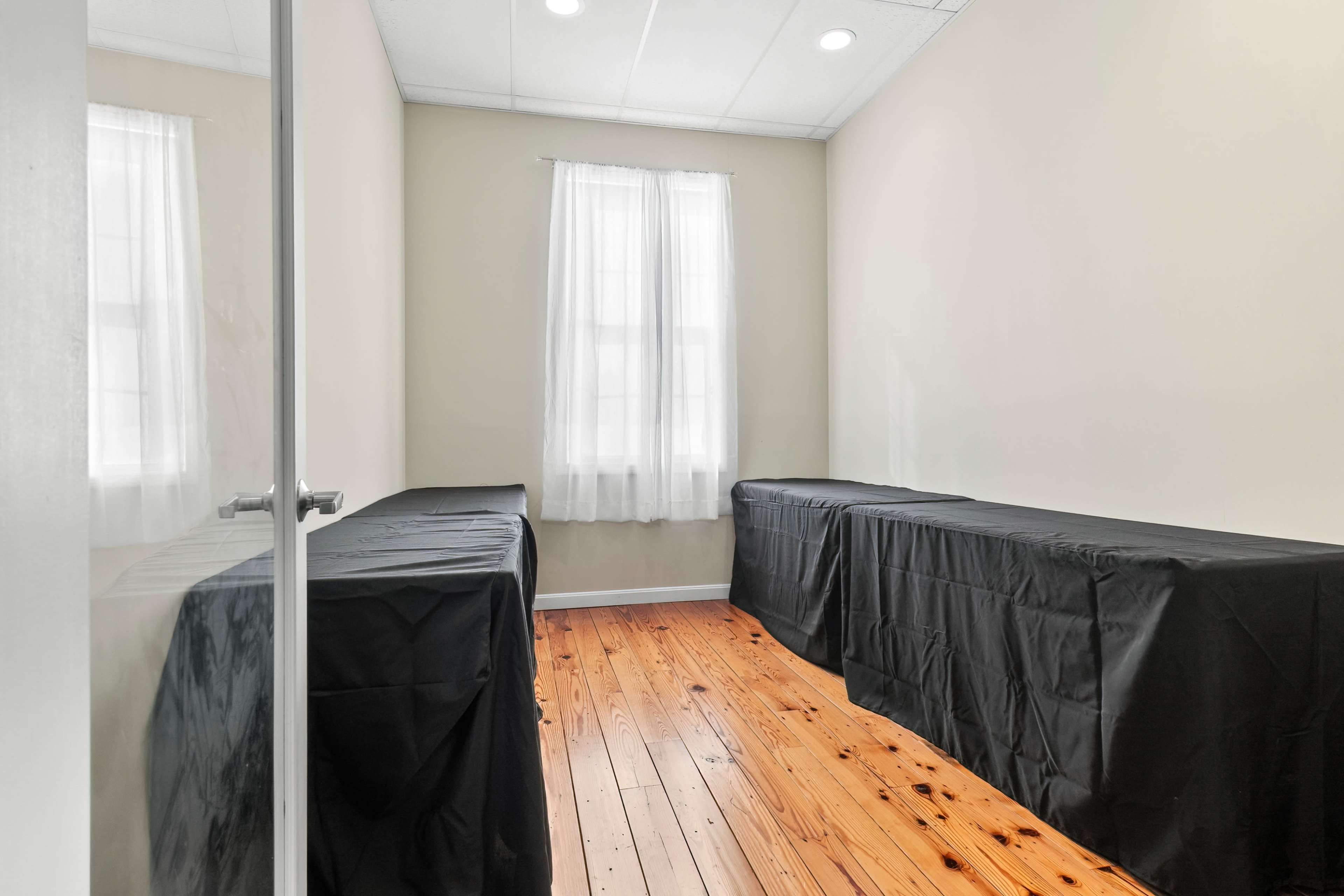 The image shows a small, empty room with wooden flooring and two tables covered in black cloth, illuminated by natural light through a window with white curtains.