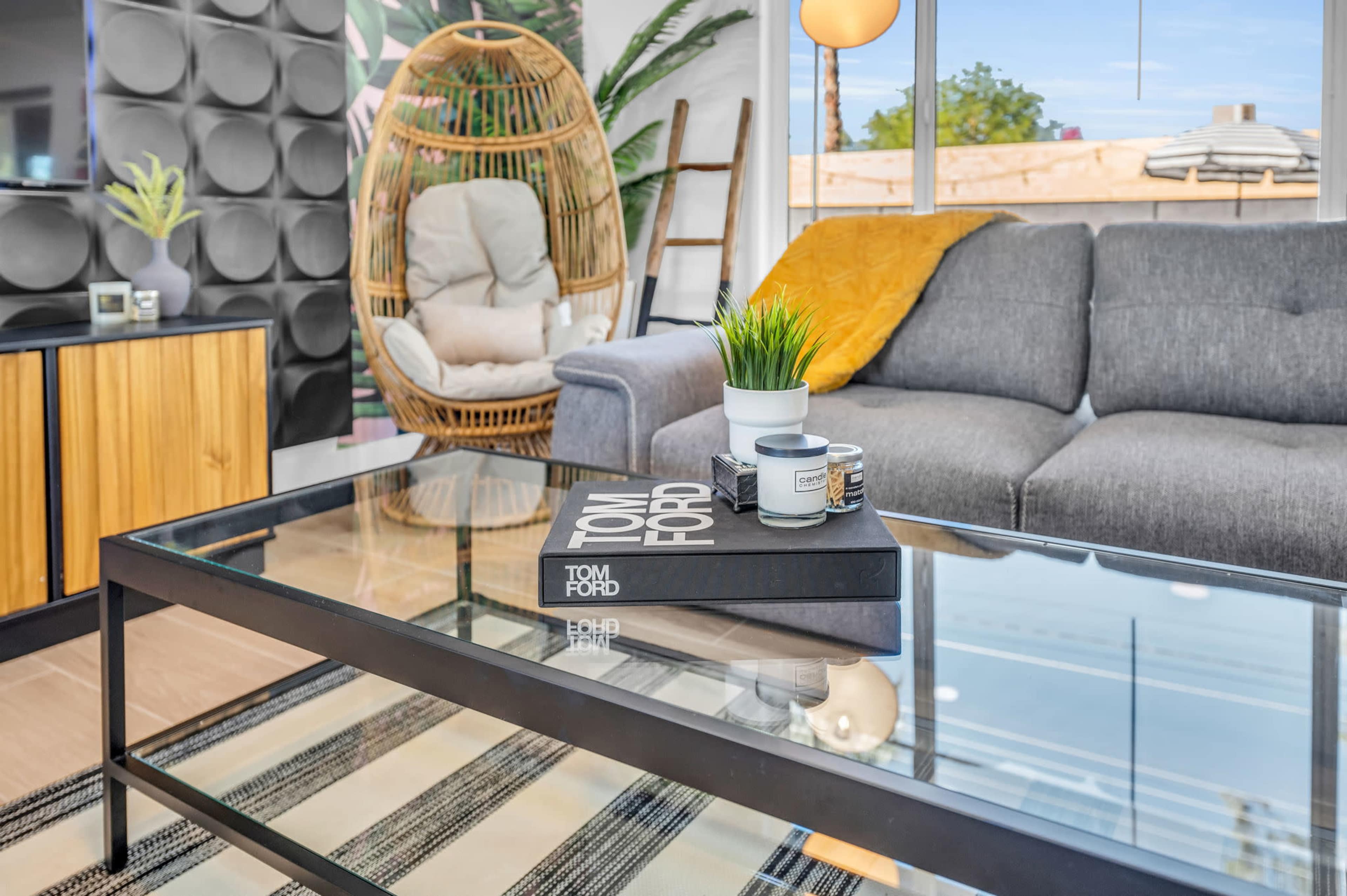 The scene features a modern living room with a glass coffee table topped with books and a decorative plant, surrounded by a gray sofa and a wicker chair.
