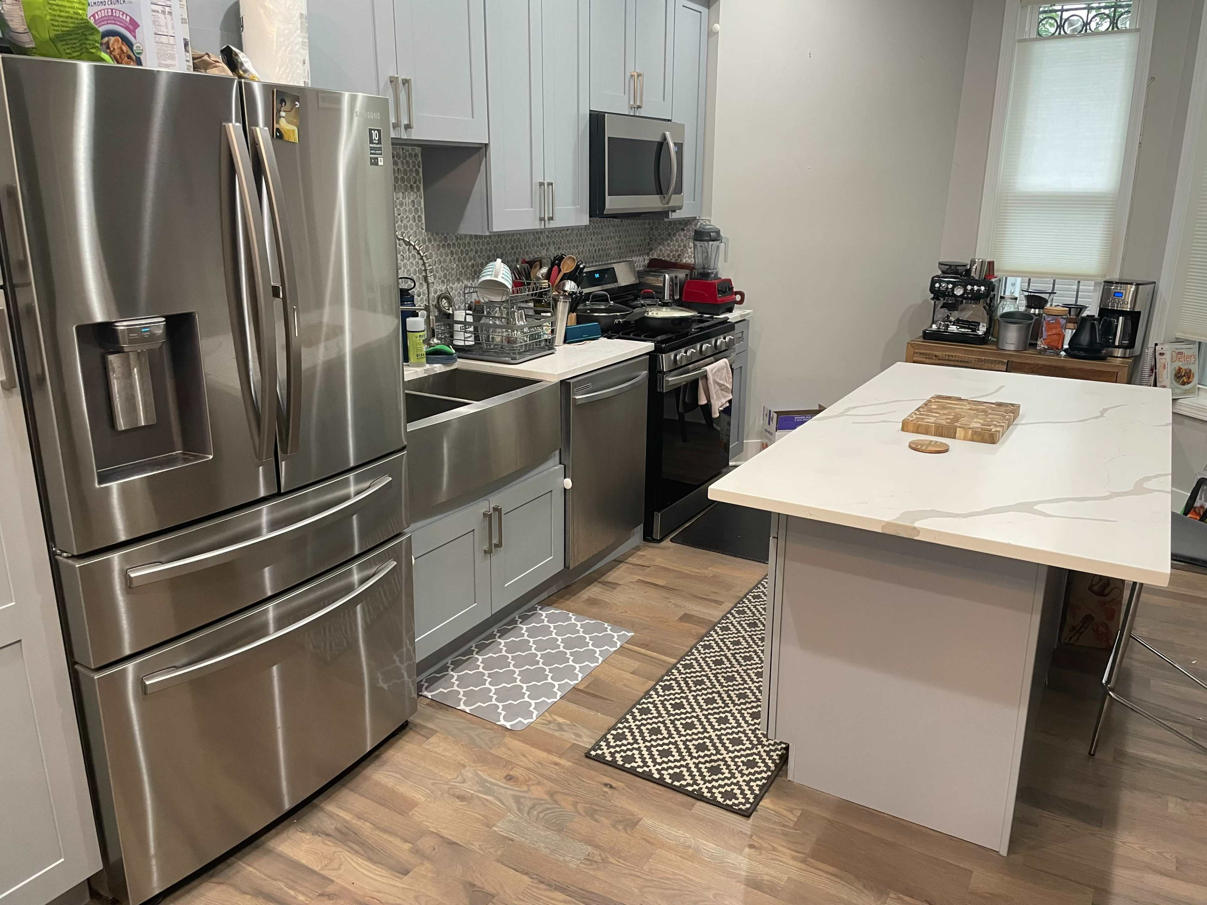 The image shows a modern kitchen featuring stainless steel appliances, a large island with a marble countertop, and a combination of light-colored cabinets and flooring.