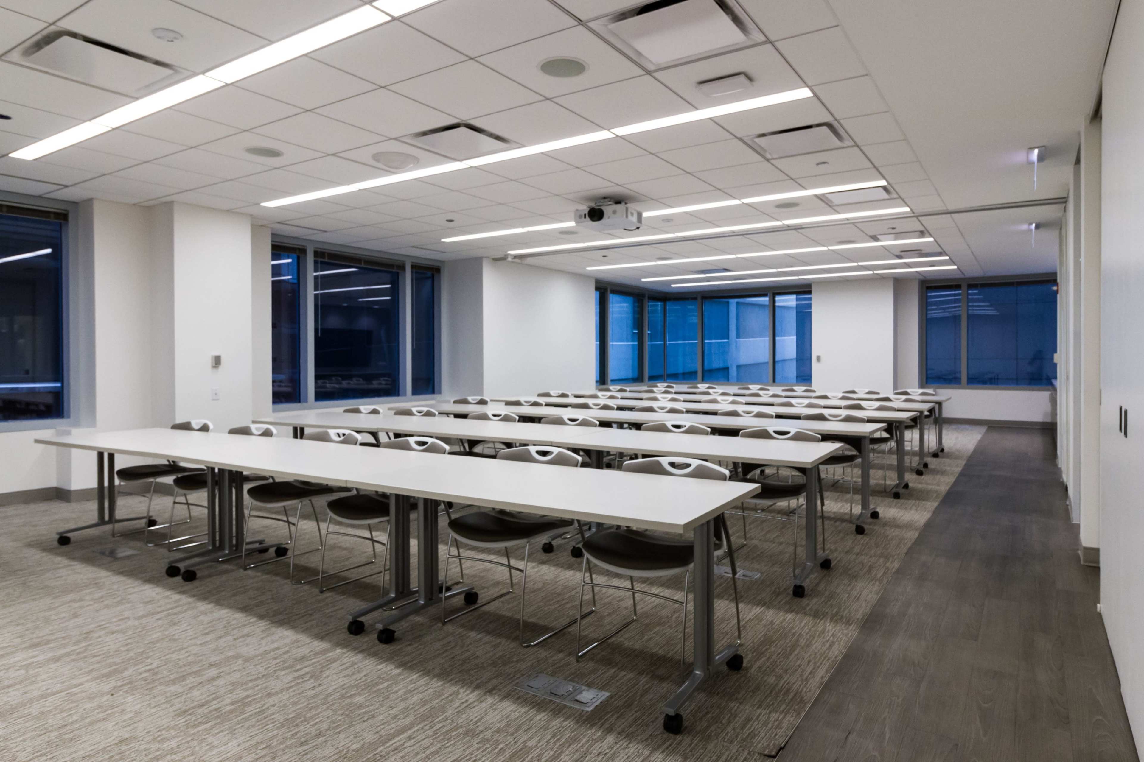 A modern classroom features rows of long tables with chairs, large windows, and overhead lighting.