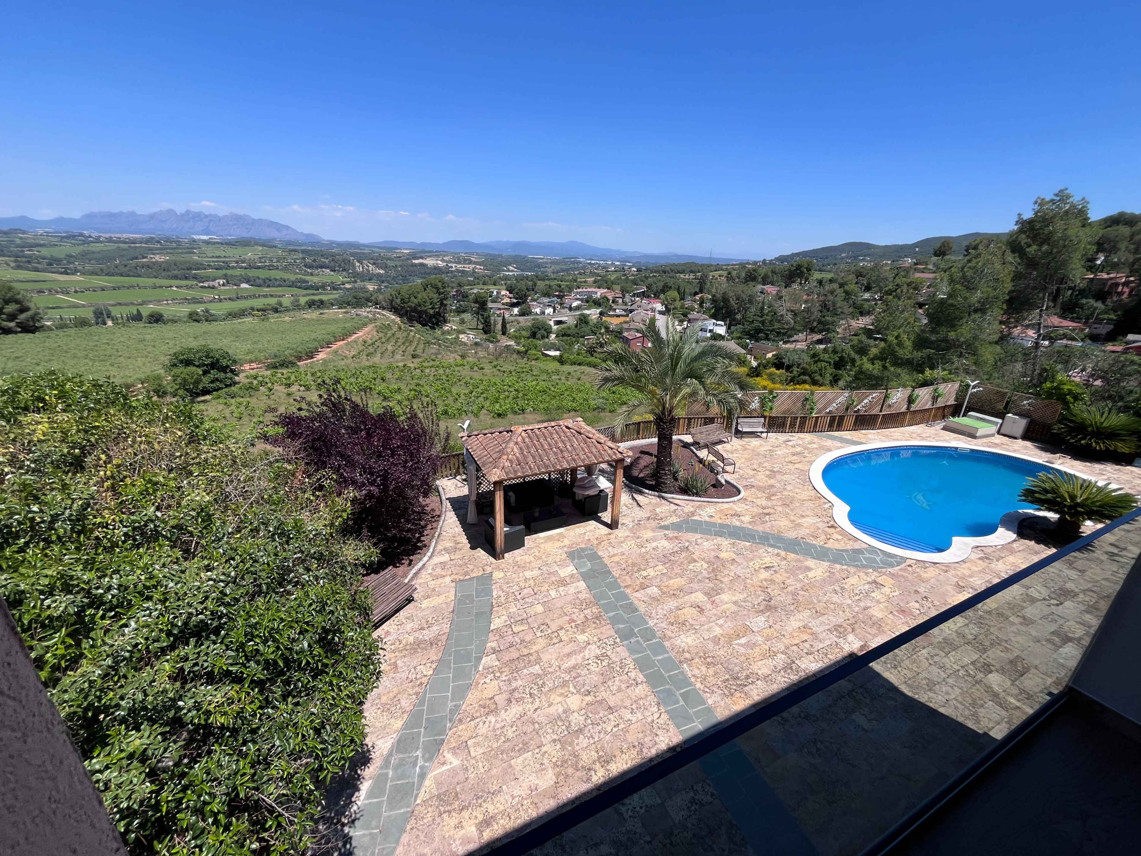 The image shows a scenic outdoor area featuring a swimming pool, a stone patio, and a view of lush green hills and vineyards in the background.