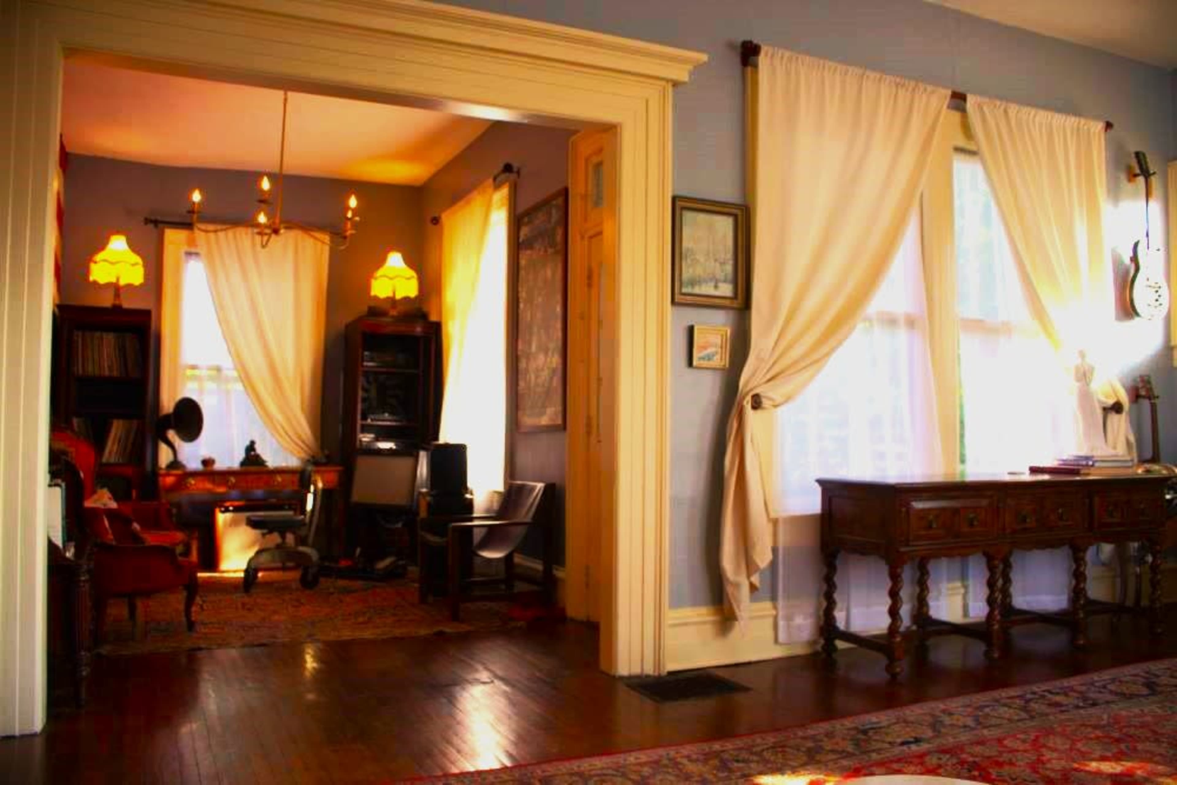 The image shows a cozy interior with two adjoining rooms, featuring large windows draped with white curtains and vintage furniture, including a console table and seating arrangements.