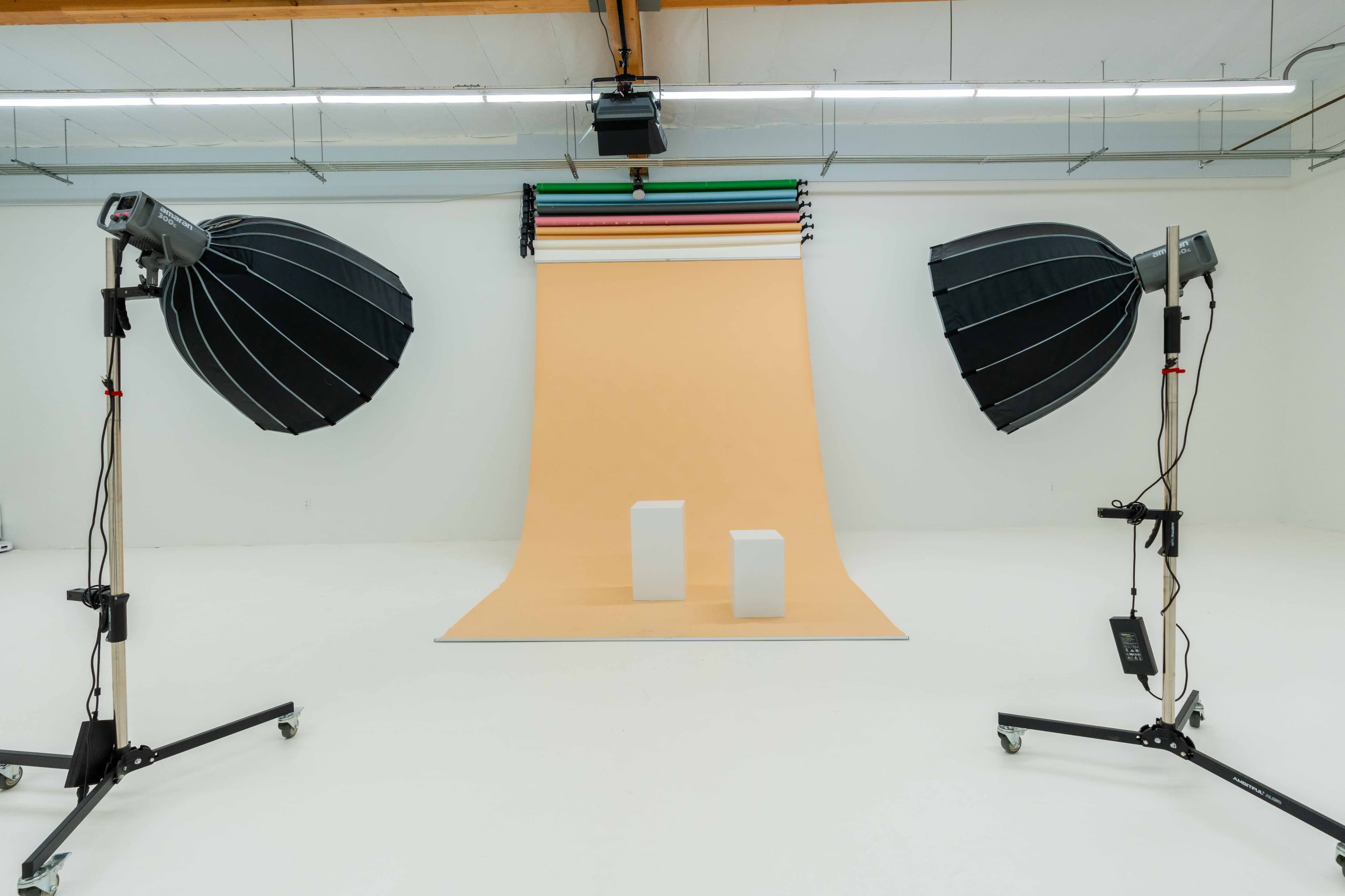 The image shows a photography studio with two large softbox lights positioned on either side of a light peach backdrop, featuring two white display cubes in the center.