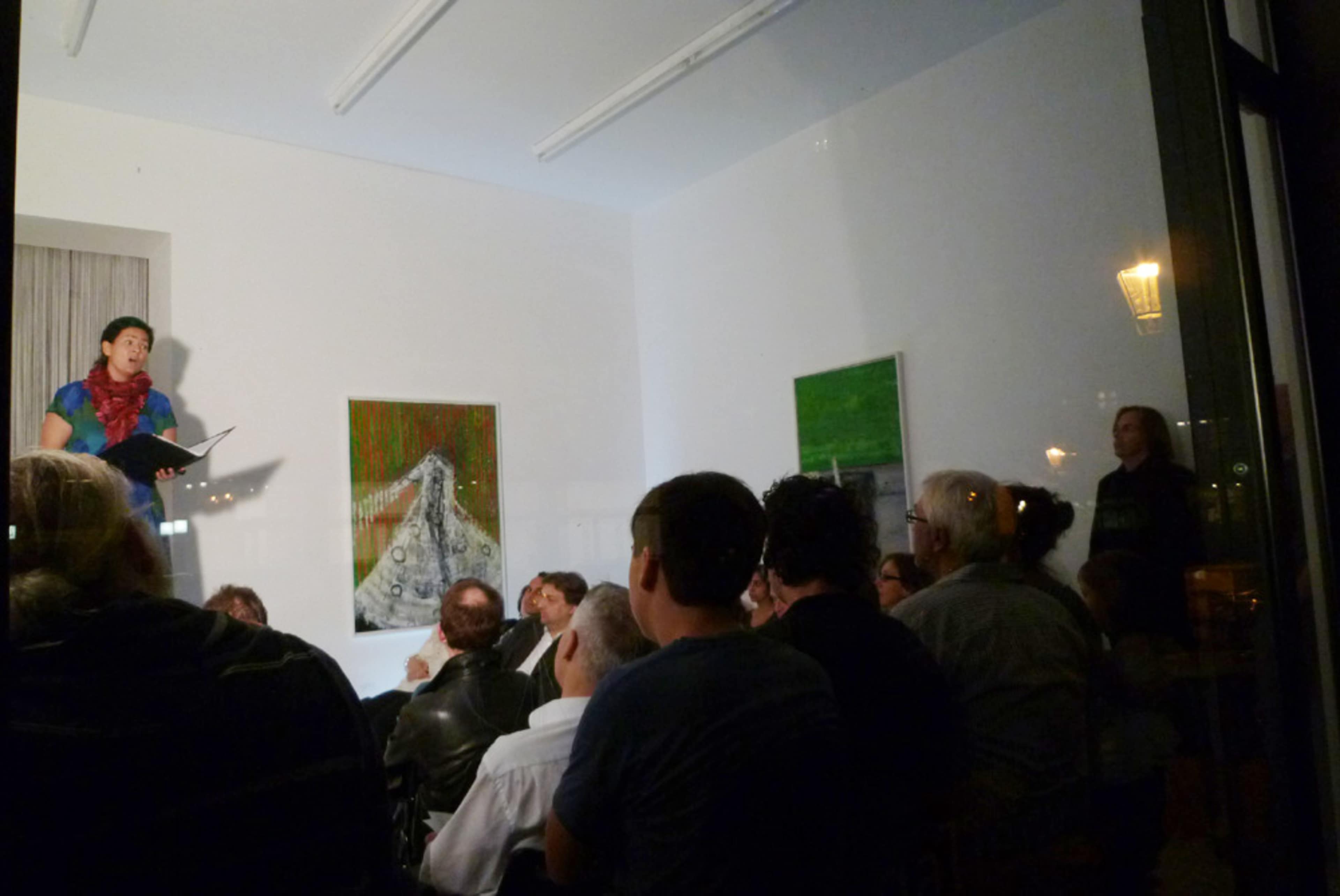 A performer stands on a stage in front of an engaged audience, with artwork displayed on the walls of a brightly lit room.