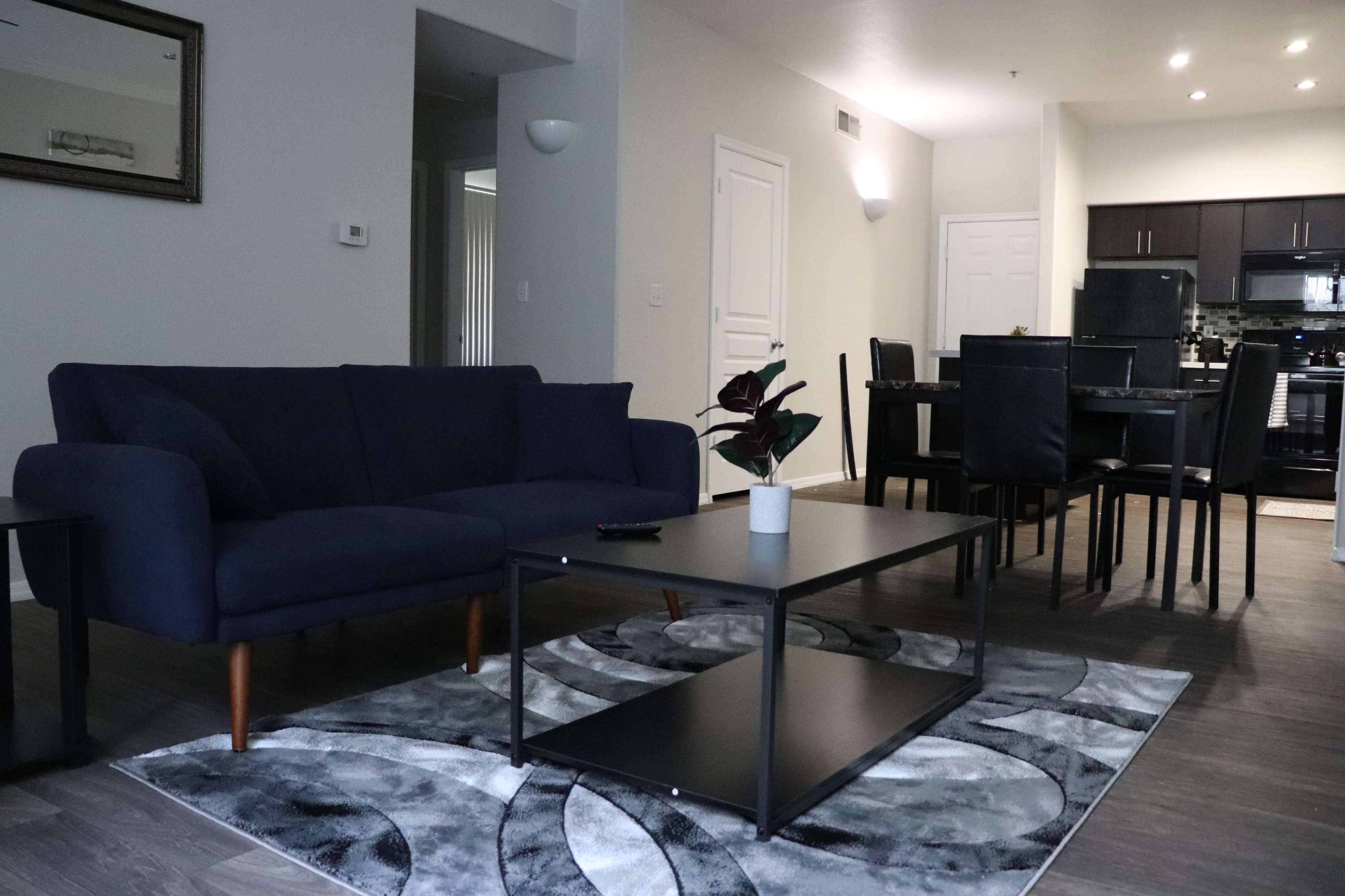 The image shows a modern living room with a blue sofa, a black coffee table on a patterned rug, and a dining area in the background.