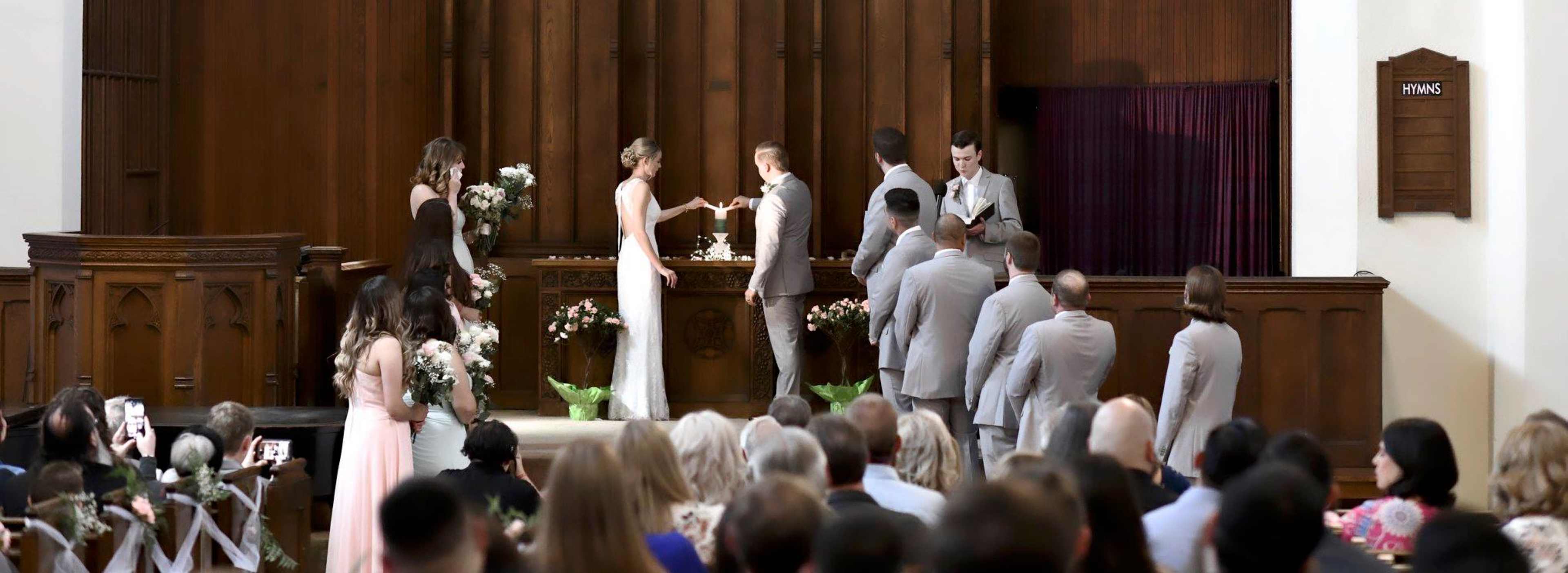 A couple exchanges vows during a wedding ceremony in a church, surrounded by their bridal party and guests.