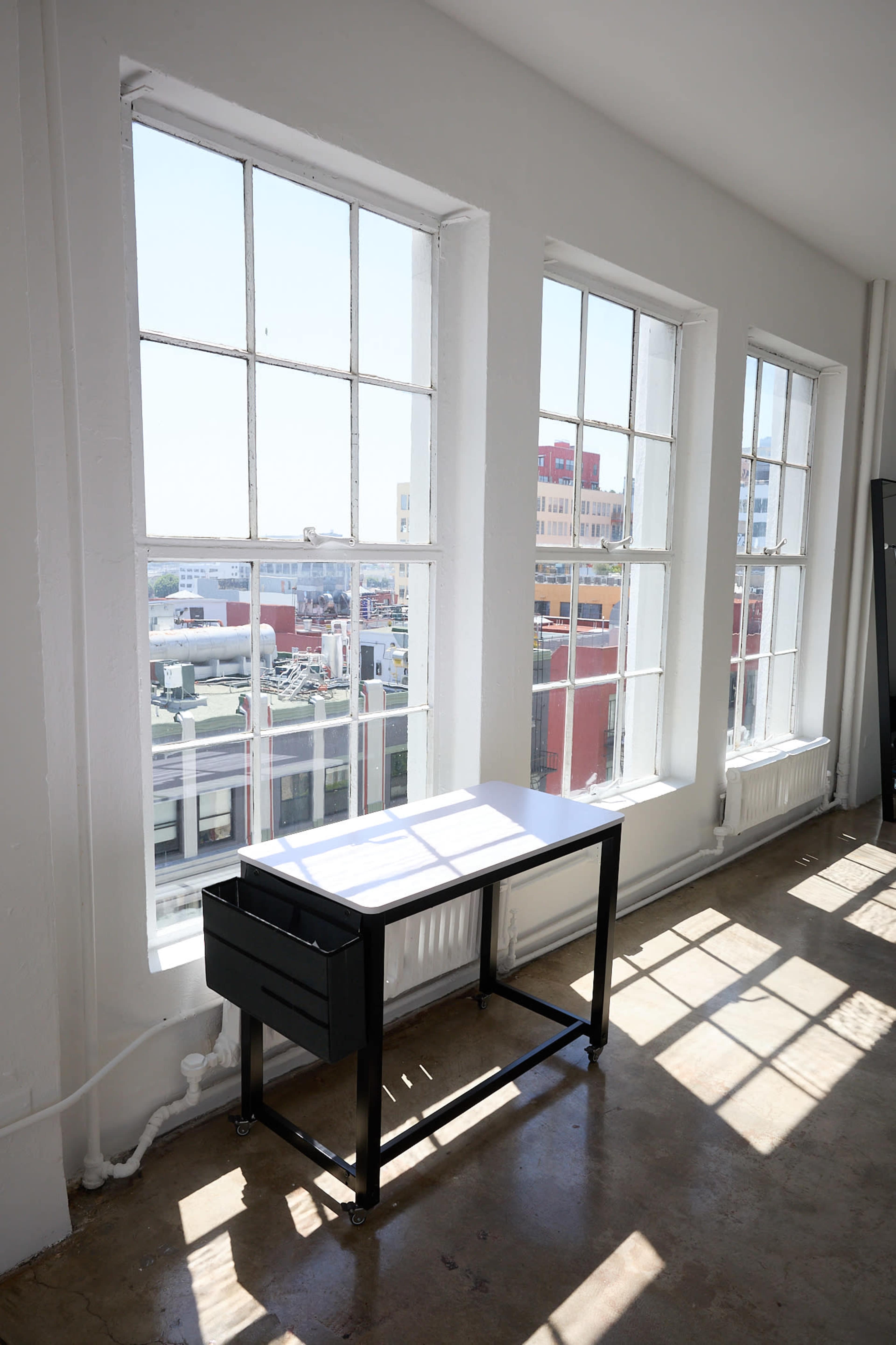 A minimalist desk stands beside large windows, allowing natural light to illuminate the space and offering a view of city rooftops.