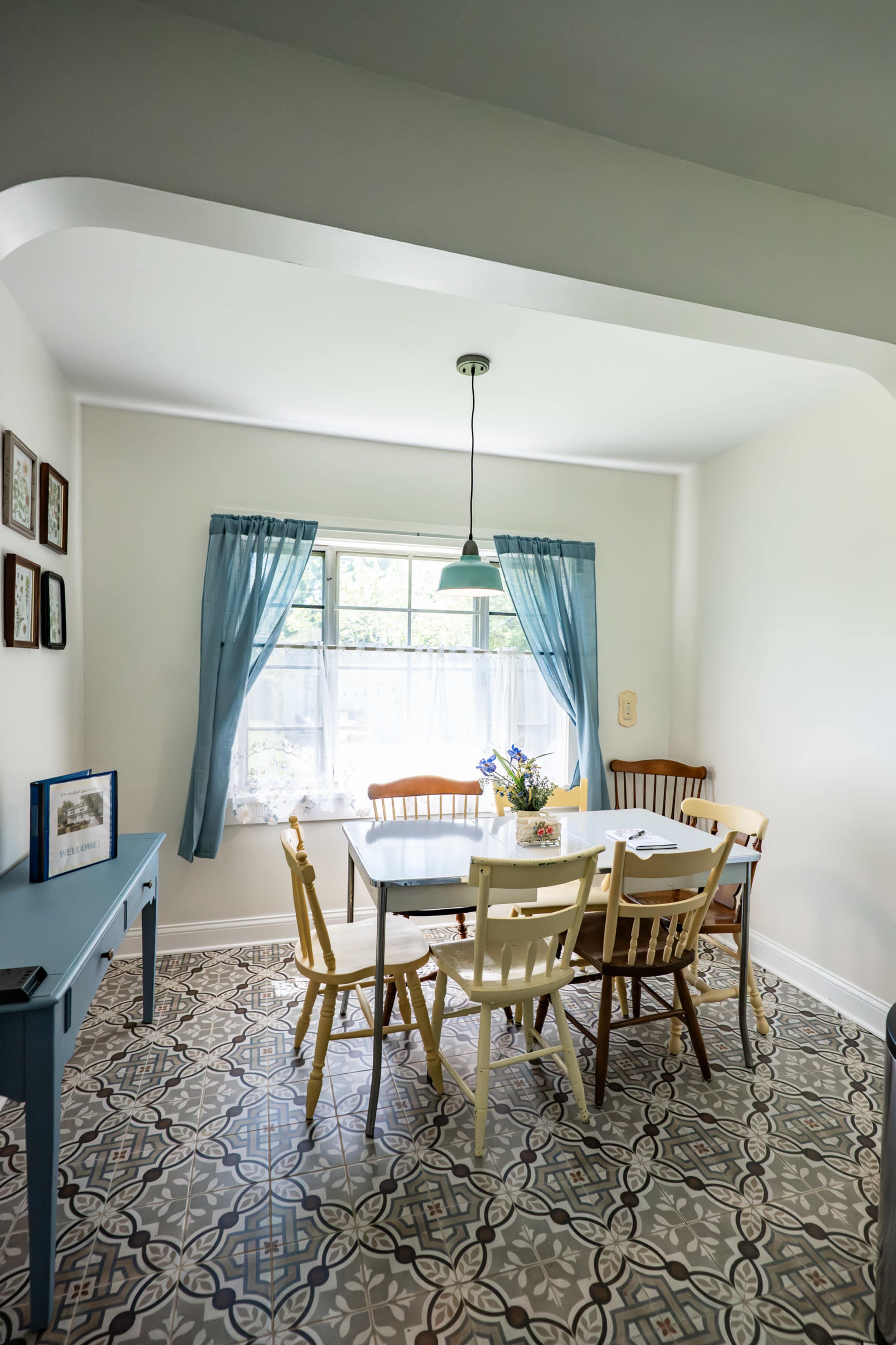 The image shows a bright dining area featuring a rectangular wooden table surrounded by chairs, with a vase of flowers on the table and large windows dressed in blue curtains.