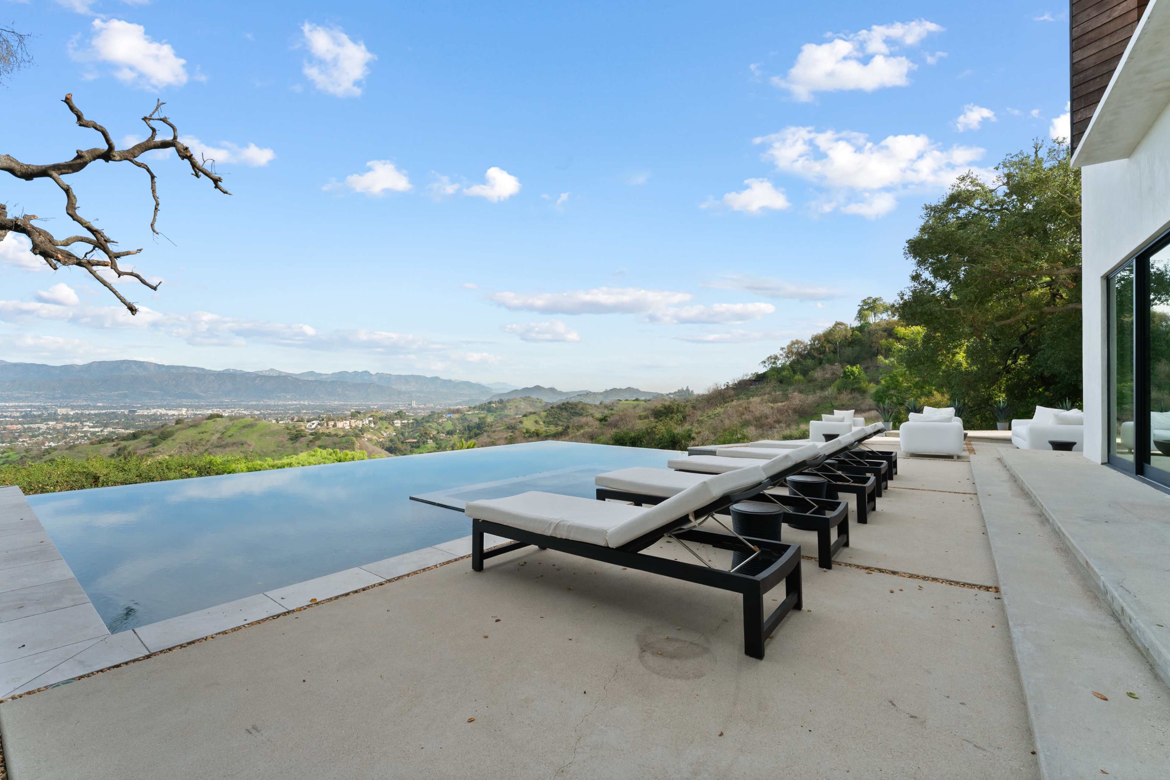 The image shows an infinity pool with lounge chairs overlooking a mountainous landscape under a clear blue sky.