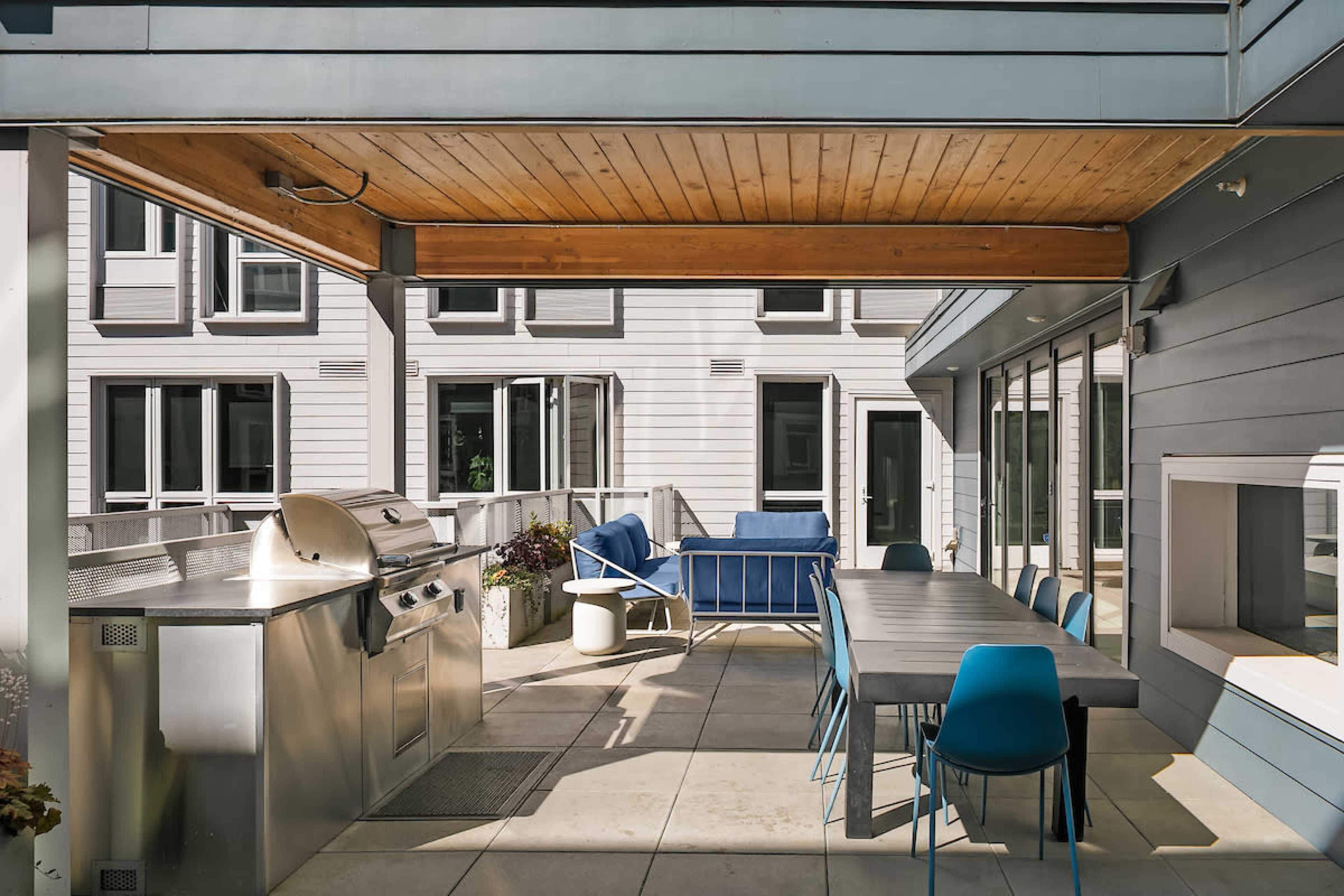 The image shows a modern outdoor patio featuring a built-in grill, a dining table with blue chairs, and a seating area with a blue couch.