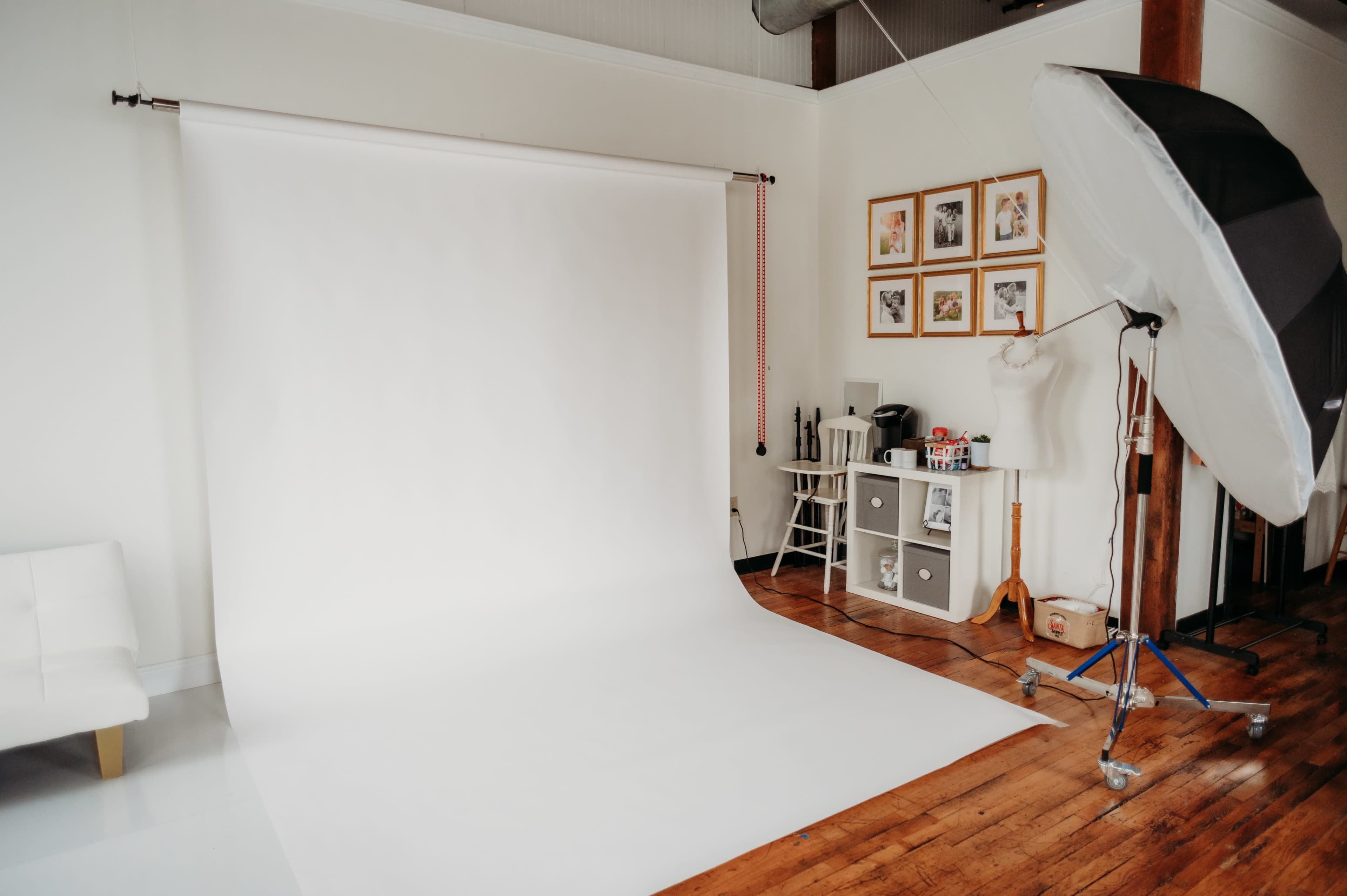 A photography studio features a white backdrop, a light source on a stand, and a small storage unit against a wooden floor.