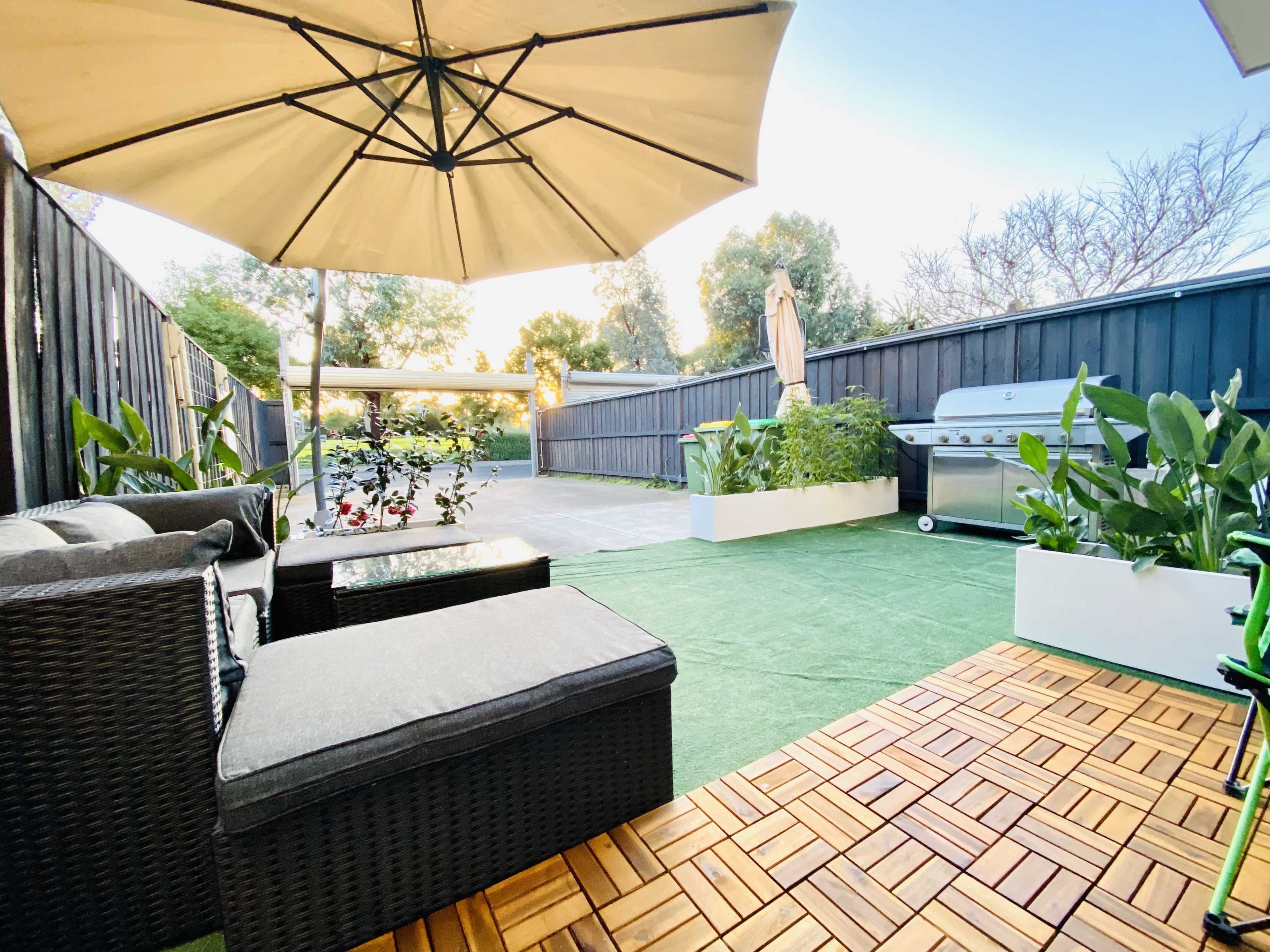 The image shows a patio area with a seating arrangement under an umbrella, artificial grass, and a barbecue grill against a wooden fence.