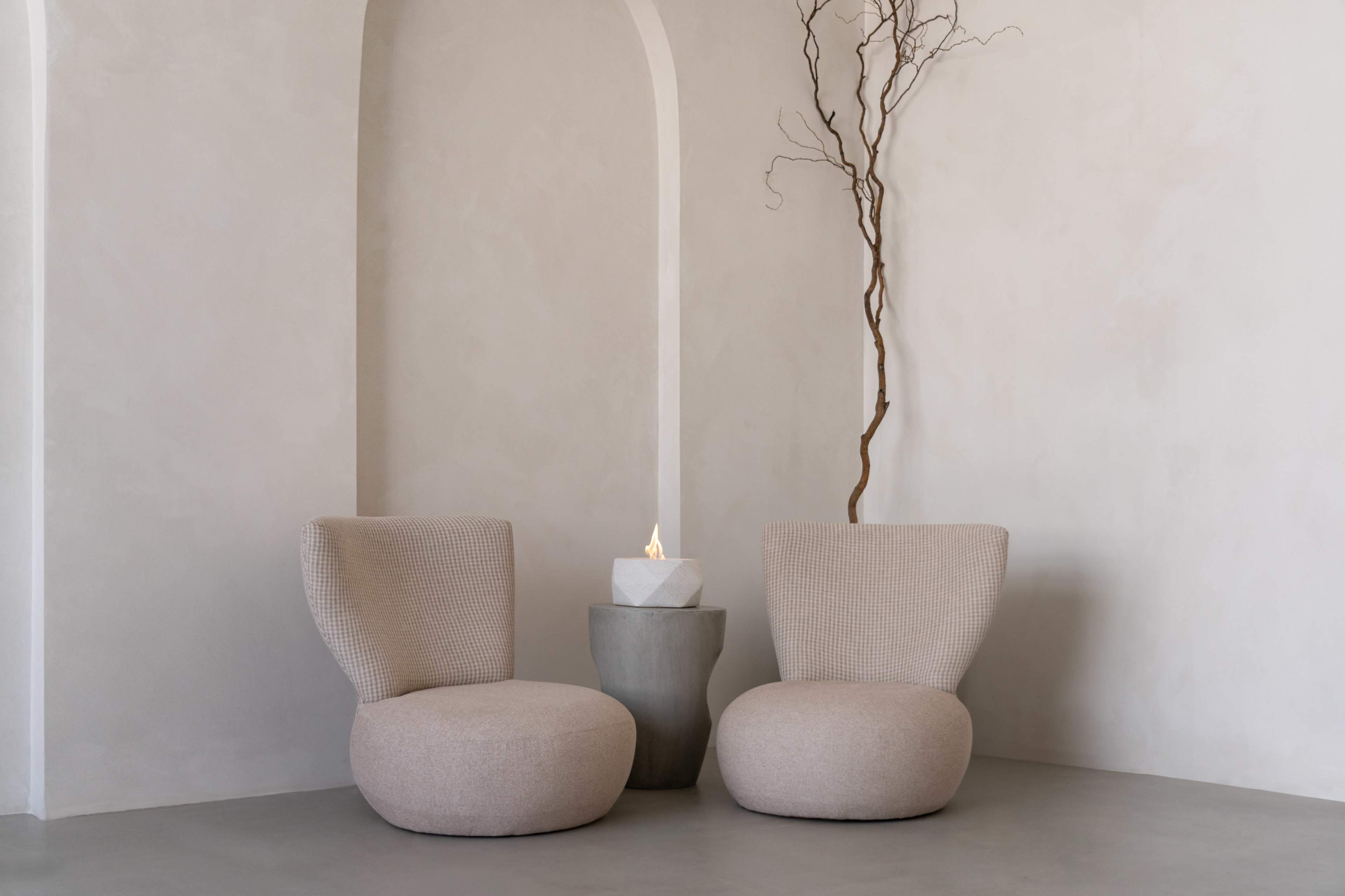 Two upholstered chairs with curved backs sit beside a stone table, which holds a small flame, against a minimalist wall with an arch.