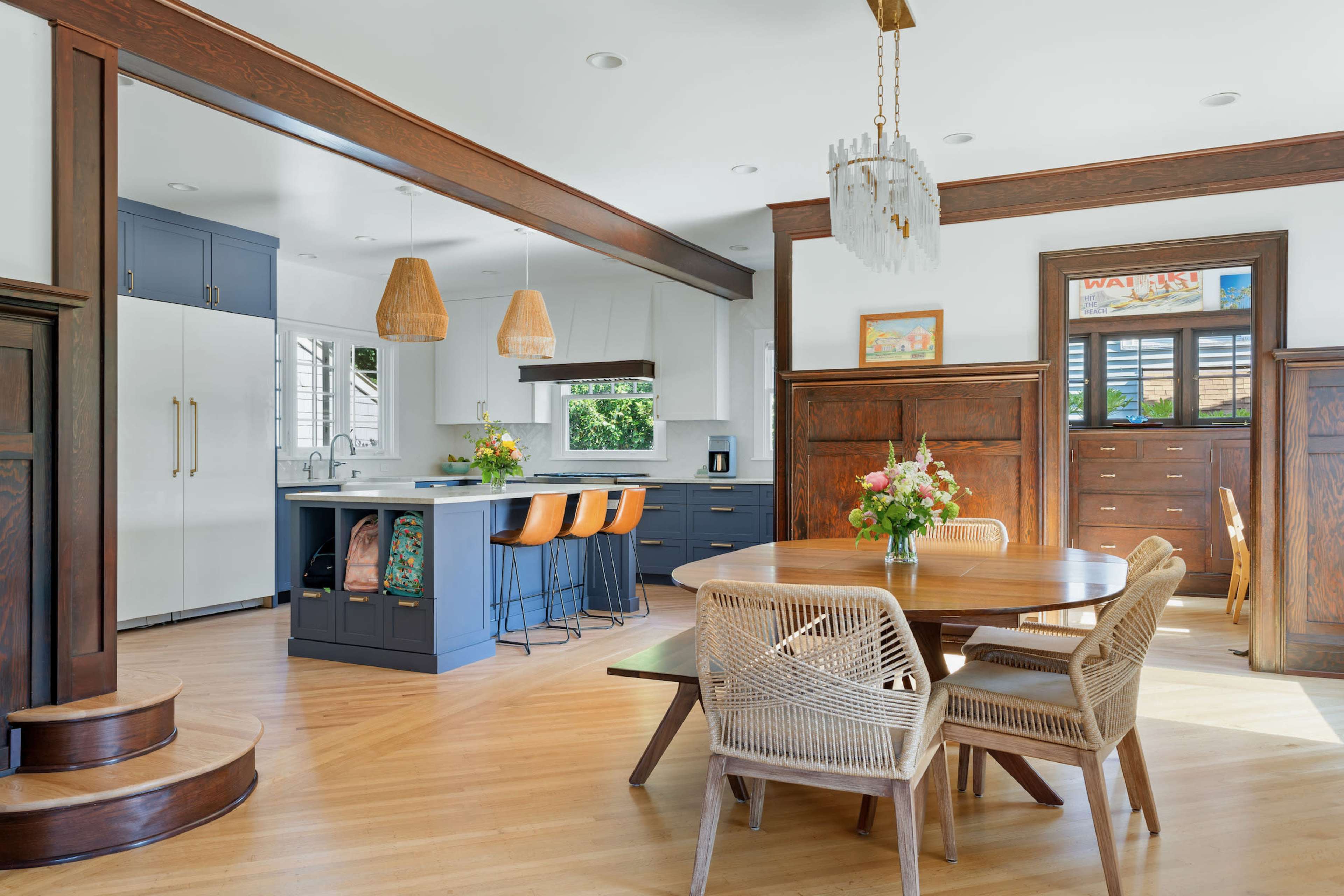 A modern kitchen and dining area featuring a round wooden table surrounded by rattan chairs, with a central island and bar stools in a well-lit space.