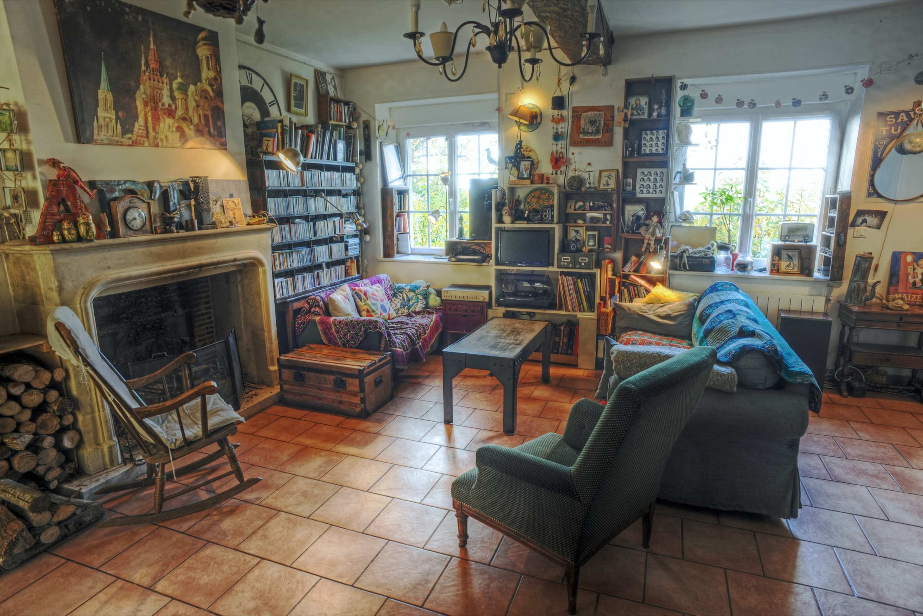 The cozy living room features a fireplace, wooden shelves filled with books, and mismatched furniture arranged around a central coffee table.